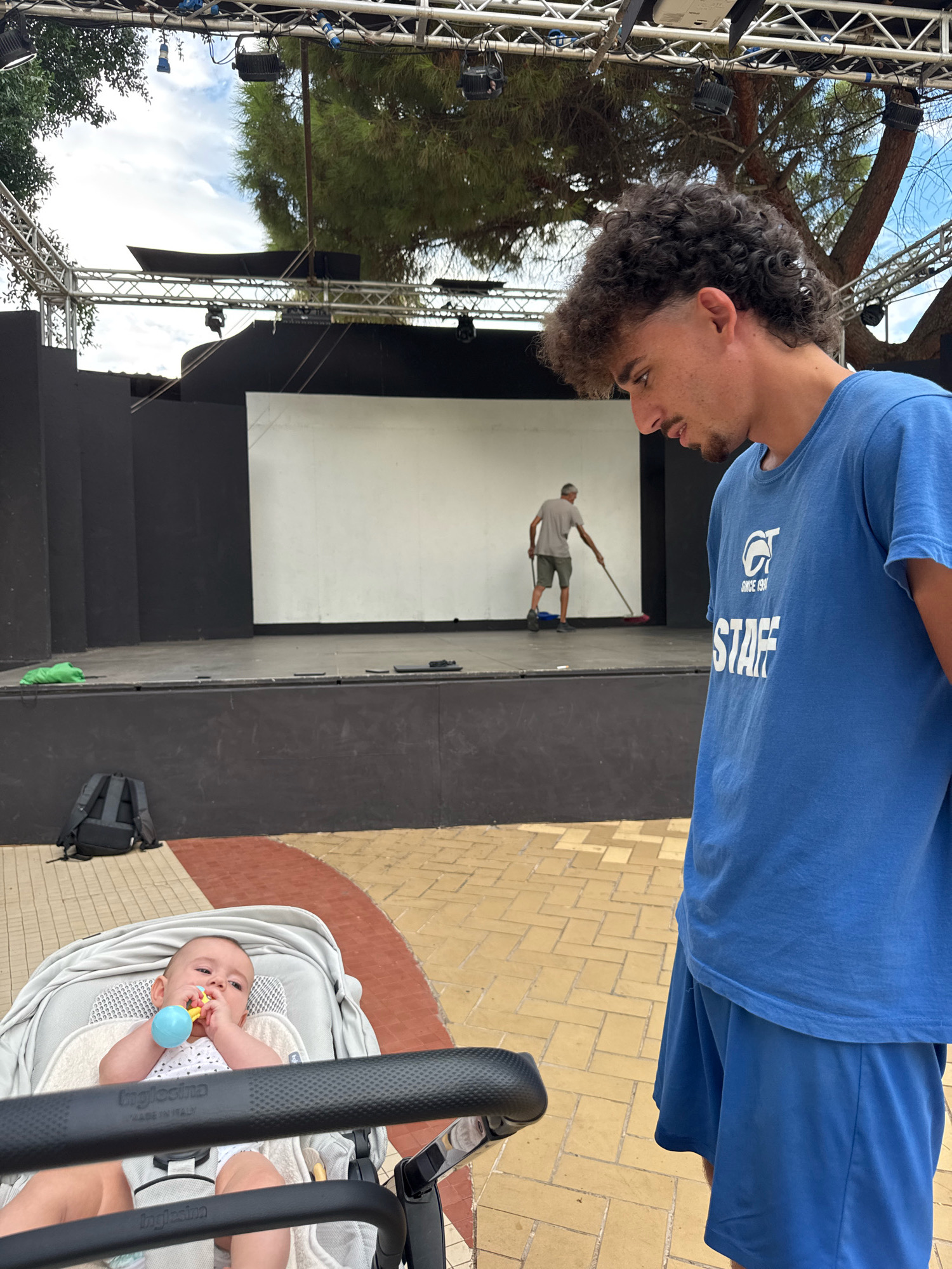 In Isola delle Femmine, Italy, a young man with curly hair, wearing a blue t-shirt with "STAFF" printed on it and blue shorts, stands in the foreground looking down and to his left. Behind him, a stage is set up with a black backdrop and a white screen. A man in a grey t-shirt and shorts is sweeping the stage. In the lower left of the image, a baby is lying in a stroller, holding a blue toy. A black backpack is on the ground next to the stage. The scene appears to be outdoors with trees and a cloudy sky visible above the stage rigging.