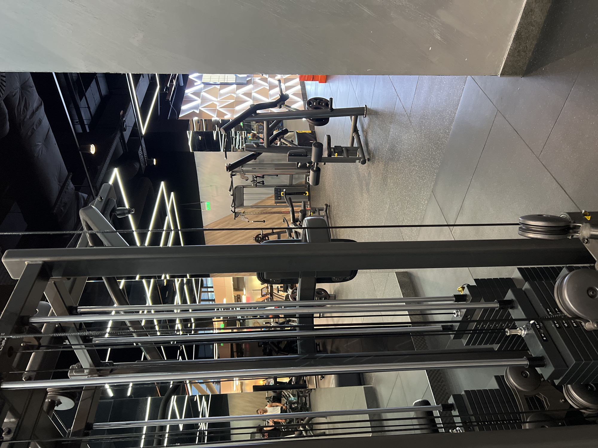 The image depicts the interior of a gym, focusing on various weight training machines. The flooring is gray and textured. The ceiling is a combination of gray and black, with some areas featuring geometric patterns of white lights.
Several weight training machines are visible, including cable machines and seated row machines. The cable machines have stacks of black weights and silver bars.
Through the reflection of the machines, people can be seen in the background.