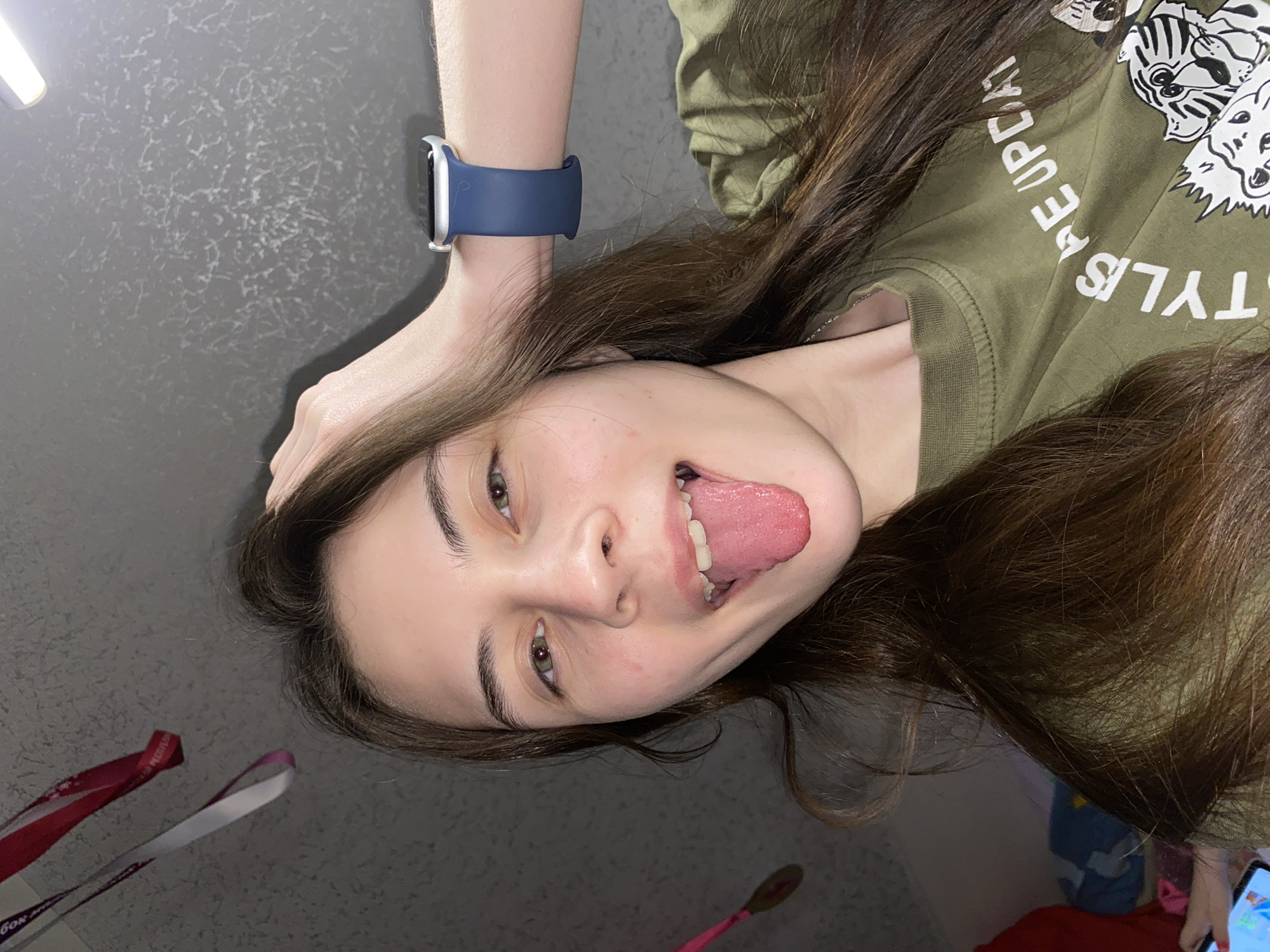 A young person, likely female, with long, dark brown hair, is captured in a playful pose, sticking out their tongue and looking directly at the camera. The image is rotated 90 degrees clockwise.
The subject is wearing an olive green t-shirt adorned with white text and graphic designs. Visible text on the shirt includes "STYLES" and "PEOPLE UPDATE" (partially visible as "PE UPDA"), along with stylized white outlines of what appear to be tiger or cat heads. On their left wrist, they wear a silver Apple Watch with a dark blue silicone band. Their complexion is fair, and their eyes are a light brown or hazel color.
The scene appears to be indoors, against a dark grey or black textured background, possibly a ceiling or wall, which features light, irregular, scrawled patterns. A bright, elongated artificial light source is visible in the upper left of the frame. In the lower left corner, two lanyards or ribbons, one red and one purple/white, are visible; the red one displays the Cyrillic letters "БОК" (BOK). The very bottom right corner shows a hint of a pink object, possibly a handle, and the edge of a digital screen.
The overall mood is light and informal, characterized by the subject's mischievous expression. The location is Votkinsk, Russia.