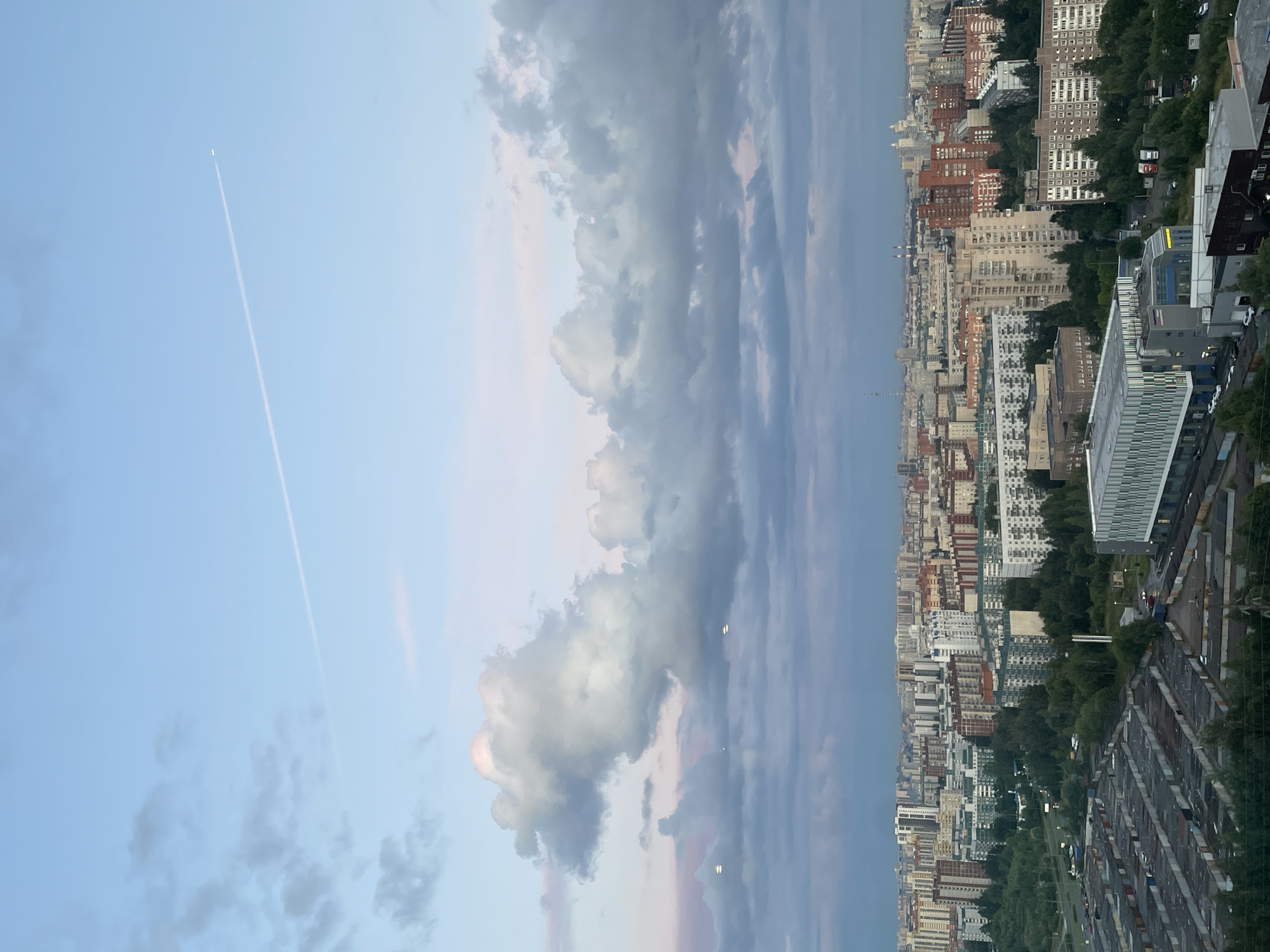 A wide-angle, eye-level outdoor shot shows a sprawling urban cityscape under a pale blue sky with scattered clouds and a contrail from an airplane. The cityscape is densely packed with a variety of residential and commercial buildings, ranging in height and architectural style. Dominating the foreground are rows of garages with corrugated metal roofs, interspersed with green trees. Beyond the garages, numerous multi-story apartment buildings are visible, some with light-colored facades and others with red brick. Further into the distance, the skyline is punctuated by taller, more modern buildings, and industrial structures. The overall impression is of a large, established city with a mix of older and newer developments. The lighting suggests it is either early morning or late evening, with soft, diffused light.