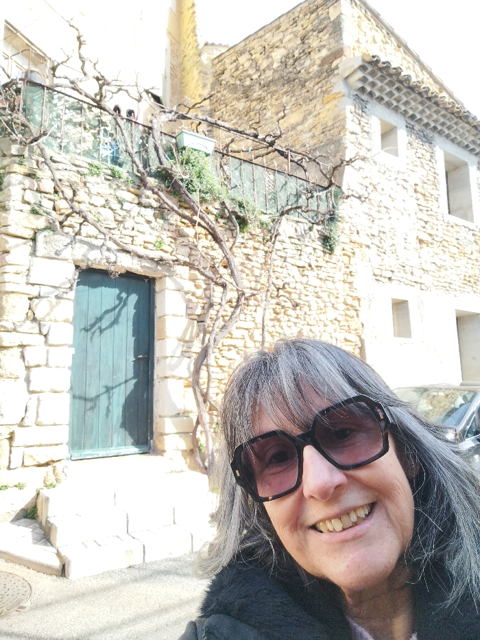 A woman with gray hair and large sunglasses poses in front of an old stone building. The building features a blue door and a balcony with a metal railing. A bare vine climbs the wall, adding to the rustic charm. A car is partially visible behind the woman, suggesting the photo was taken on a street. The lighting suggests a sunny day, casting shadows on the door and building.