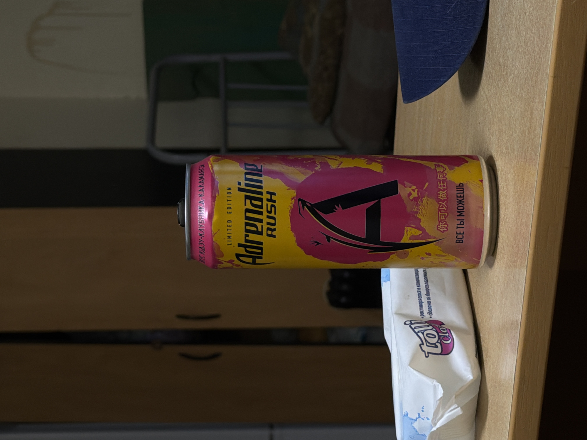 A medium shot captures a can of "Adrenaline Rush" energy drink, emblazoned with "LIMITED EDITION" and the brand's signature "A" logo, positioned on a wooden table. The can is predominantly yellow and pink, with splashes of color and black lettering. To the left of the can lies a white package of "Toll" brand wet wipes, featuring blue accents and text in Russian. The background is dimly lit and out of focus, suggesting an indoor setting with furniture, possibly a dormitory or a simple room. The overall scene appears to be a still life, with no human presence or activity evident. The image does not contain information that would allow for its specific location in Rostov-on-Don, Russia.
