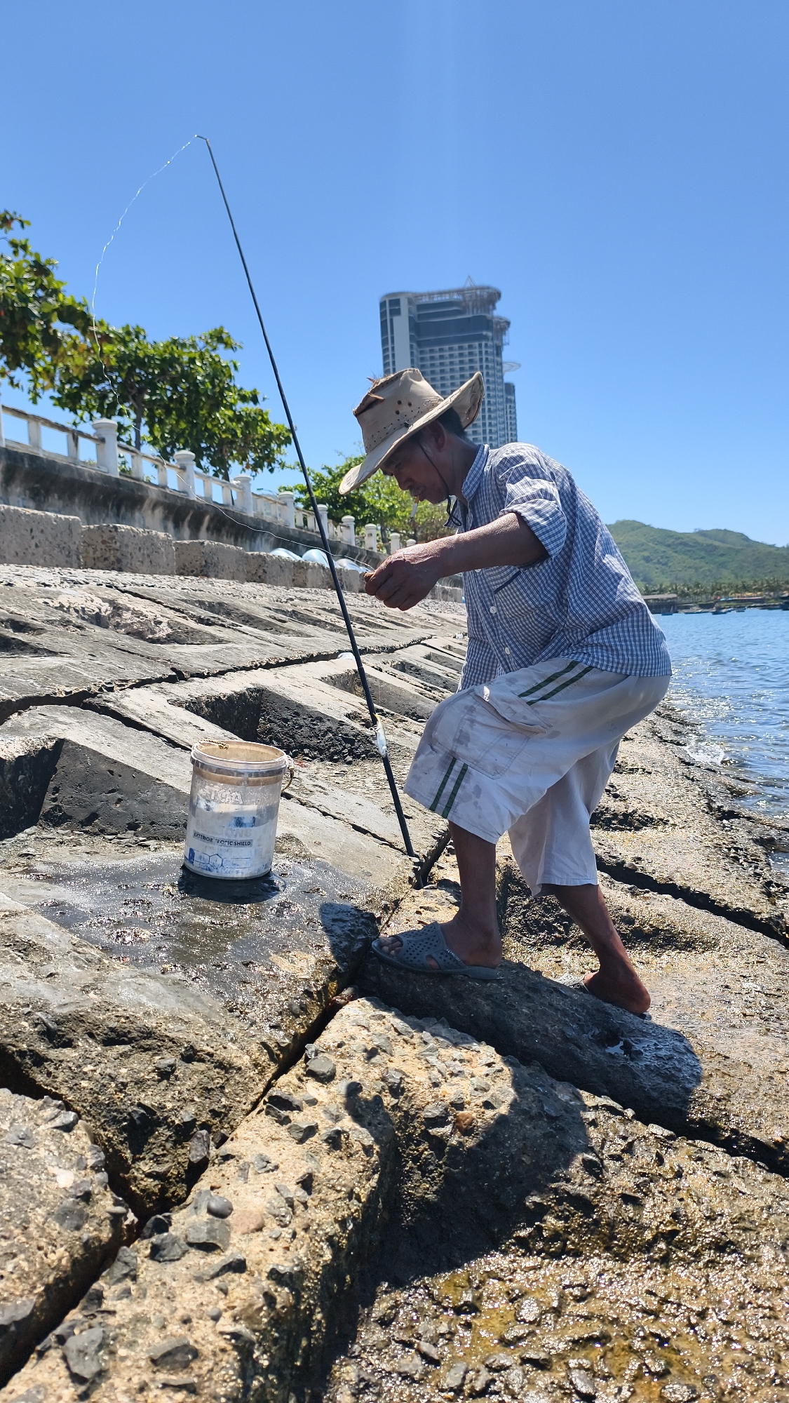 A man wearing a hat and a checkered shirt is fishing on a rocky shore in Nha Trang Ward, Vietnam. He is holding a fishing rod and appears to be baiting his hook. Next to him is a bucket. The background shows a modern building, trees, and the sea. The day is clear and sunny.