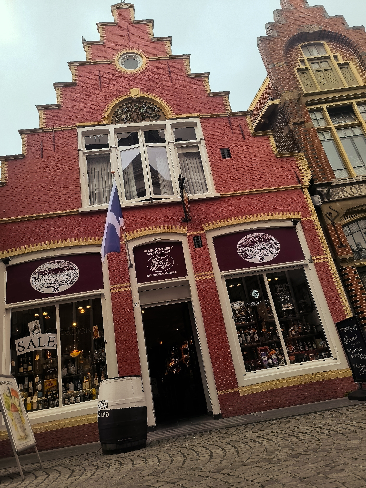 This is a medium-angle, low-light, eye-level outdoor shot of a red brick building in Hoorn, Netherlands, specifically on the Rode Steen square. The building features a distinctive stepped gable with yellow decorative trim. The facade is predominantly red brick, with white window frames and a white entrance. A blue and white flag hangs from a flagpole on the left side of the building.
The building's ground floor has two large storefront windows. The left window displays a "SALE" sign, a poster featuring a drink, and shelves filled with bottles. The right window is the entrance to a shop labeled "WIJN & WHISKY SPECIALZAAK" (Wine & Whisky Specialty Store) with the text "BESTE SLIJTERIJ VAN NEDERLAND" (Best liquor store in the Netherlands) below. Inside the shop, numerous bottles of alcohol are visible.
In front of the entrance, a black and white barrel with the words "SOMETHING NEW WE DID" is placed on the cobblestone street. To the left of the barrel and closer to the building is an A-frame sign with a poster of a drink. A black chalkboard sign is visible on the right side of the image. The cobblestone pavement is characteristic of older European city streets. The sky is overcast.