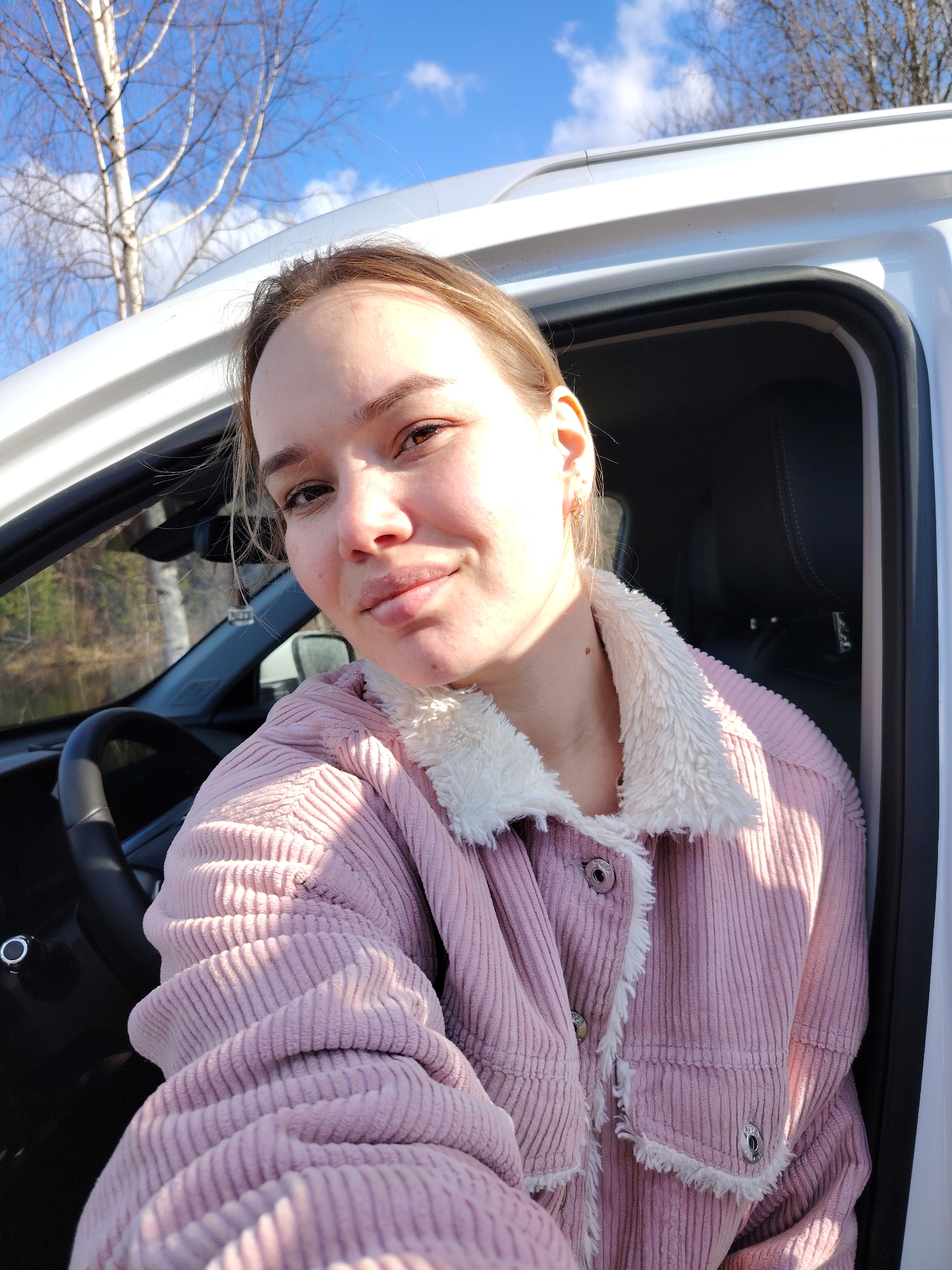 A close-up selfie shows a young woman seated in the driver's seat of a white car, outdoors in what appears to be Chaikovsky, Russia.
The woman has light brown hair, pulled back, and light eyes. She looks directly at the camera with a content, slight smile. Her face is illuminated by bright sunlight, casting some shadows. She is wearing a light pink, corduroy jacket with a white, fluffy sherpa collar and cuffs, fastened with metal buttons. A small, subtle earring is visible in her right ear, and a mole is present on her neck.
The interior of the car is dark, with the steering wheel visible on the left, indicating a left-hand drive vehicle. The car's white roofline and roof rails are prominent against the sky. The side door is open, revealing a black rubber seal.
The background shows a clear blue sky with scattered white clouds. A distinct, bare tree, likely a birch with its light bark, stands prominently. Further in the background, through the car window, more bare trees and forest-like vegetation are visible, suggesting a natural setting such as woods or a park.
The lighting indicates bright daytime, likely mid-morning or afternoon. No visible text is present in the image. The overall mood is relaxed and casual.