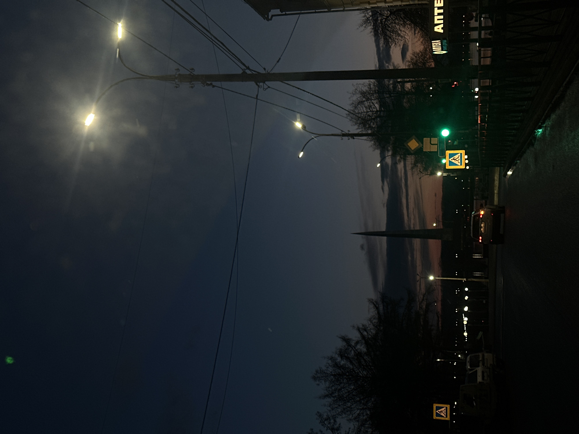 This media file captures an urban street scene at dusk or night, oriented as if viewed looking down a street. The sky is a deep blue-grey, with faint traces of orange and pink light visible on the horizon, suggesting late evening.
In the foreground and stretching diagonally across the left side of the frame is a prominent utility pole adorned with numerous overhead power lines. Several bright streetlights emit strong glows and light halos, illuminating the foreground and extending into the distance, creating a sense of depth along the street.
To the right, a road is visible, with a single car driving away, its taillights appearing as red dots. A green traffic light hangs above the street, and two distinct yellow diamond-shaped pedestrian crossing signs, each featuring a black border and a white pedestrian symbol, are visible. One is in the mid-ground, and another is closer to the bottom right of the frame.
Further back and along the right edge of the street, the dark silhouettes of bare trees are visible against the twilight sky. A building sign with the Russian word "АПТЕКА" (Pharmacy) is illuminated in the upper right corner, serving as a clear location cue. In the distant background, a tall, dark, and slender pointed structure, possibly a monument or a spire, stands against the fading sky. Numerous smaller, glowing points of light from distant streetlights dot the horizon.
No people are visible, contributing to a quiet, still, and somewhat tranquil atmosphere. The visible text and general urban infrastructure are consistent with a setting in Votkinsk, Russia.
