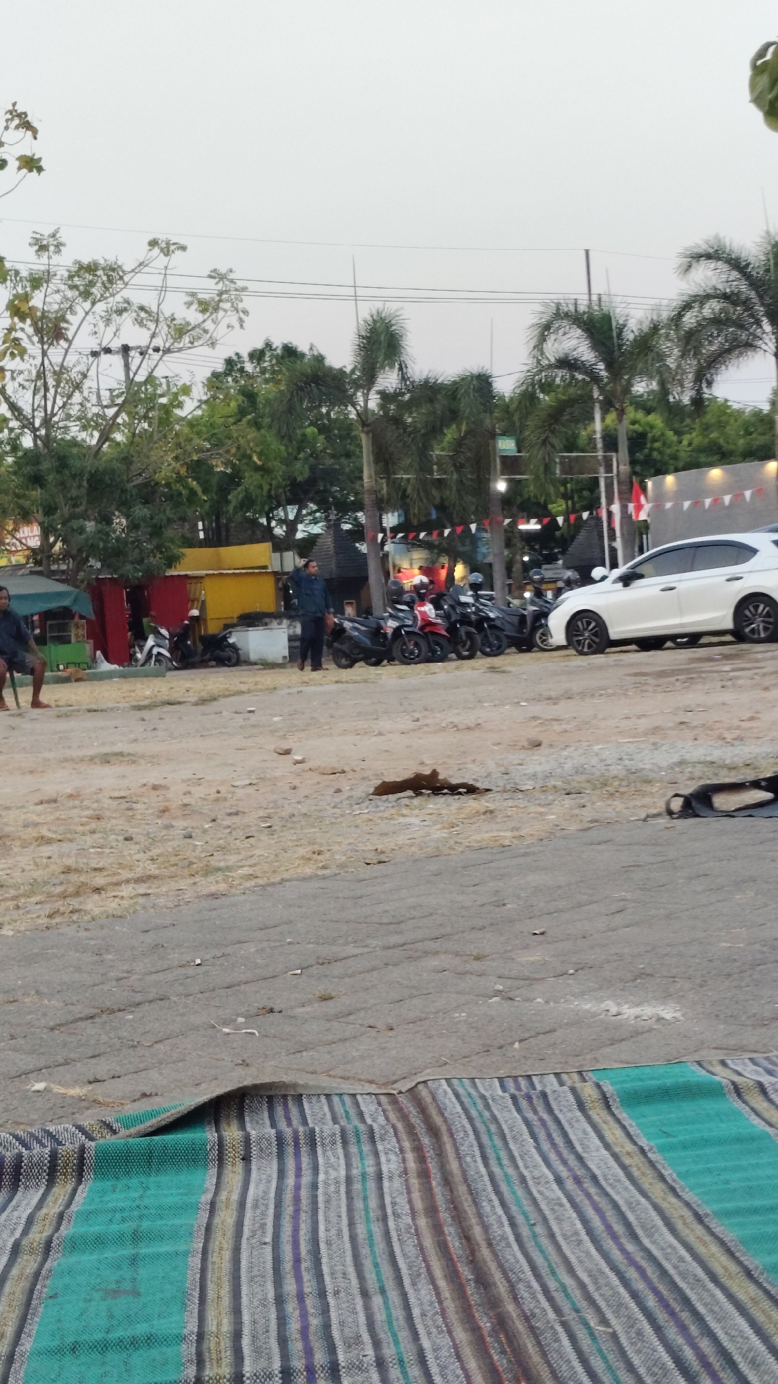 A wide shot captures a scene in Madiun, Indonesia, likely near a public gathering or recreational area. In the foreground, a colorful, striped mat is laid out on paved ground. The middle ground reveals a sandy, dirt area with sparse dry grass, where several motorbikes are parked. A white car is visible to the right, parked near a building adorned with red and white triangular flags. Behind the motorbikes, a row of food stalls with colorful facades, including yellow and red containers, are set up. A man in a dark blue shirt and pants stands near the stalls, looking towards the left. Another person, dressed in dark clothing, sits on a chair to the far left, facing away from the camera. Palm trees and other lush green foliage form a backdrop, suggesting a warm climate. The sky is overcast, casting a soft, diffused light over the entire scene. The overall impression is one of an outdoor marketplace or a casual gathering spot in a tropical urban environment.