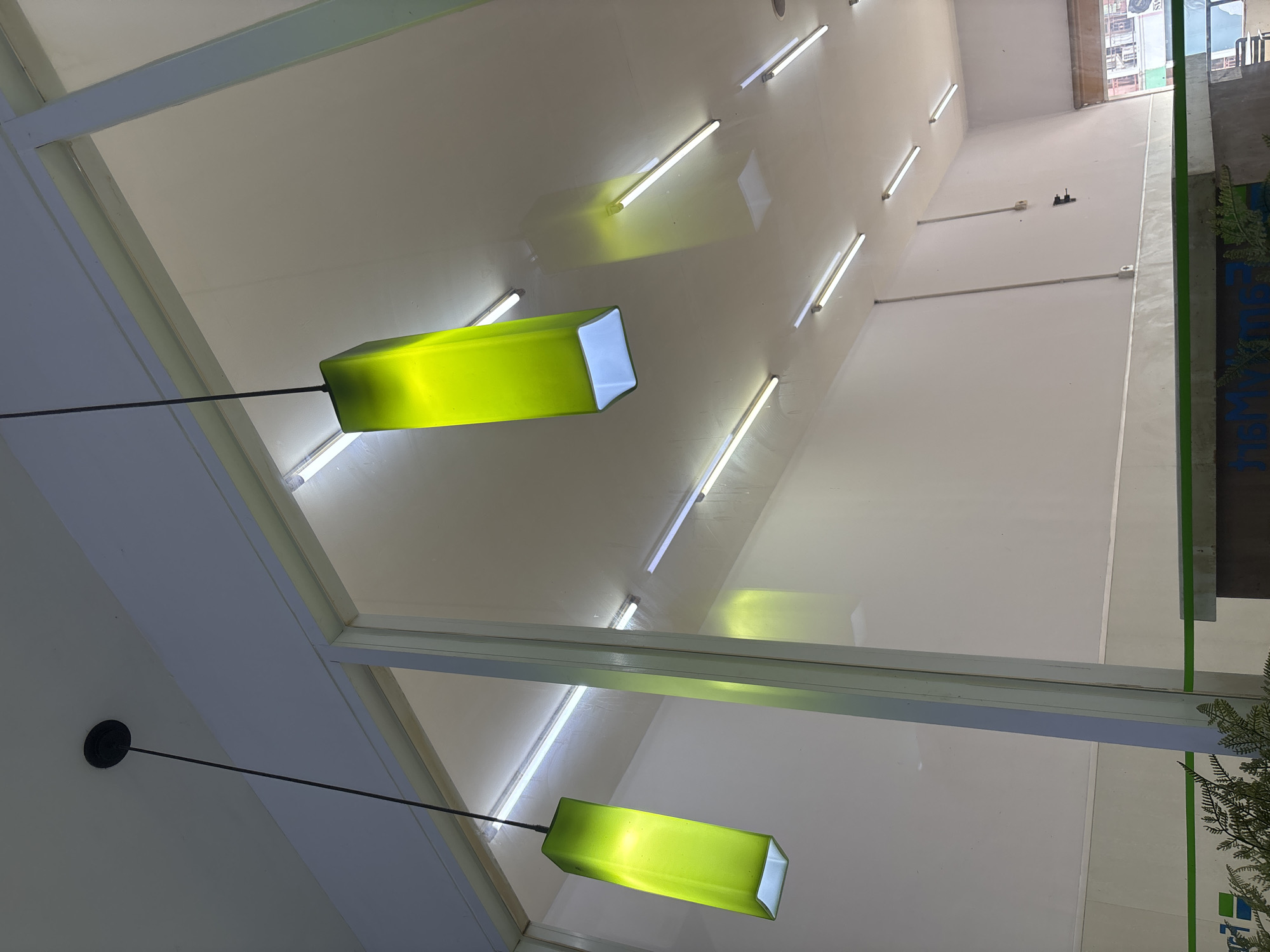The image captures an upward-looking perspective within a modern interior space, likely a commercial building in West Jakarta, Indonesia. Two prominent rectangular pendant lights, glowing vibrantly in lime green, hang from the ceiling on slender black rods. The upper pendant light is closer to the viewer and angled slightly, while the lower one is further away and more central in the frame.
The ceiling is a light off-white or pale gray, featuring numerous parallel fluorescent tube lights or LED strips. These lights emit a cool white glow and create reflections on the ceiling surface, particularly around the green pendant lights. A large glass partition or window runs horizontally across the mid-section of the image, reflecting the interior lights and creating a sense of layered space. This glass structure extends vertically along the right side of the frame, showing more of the building's interior and possibly an exterior view or another room through reflections.
On the far right, a vertical section of a bright green wall or panel is visible. Below it, part of a dark blue sign with white capital letters can be seen, spelling out "PARMA" followed by a partial, geometric green logo. Further back, along a distant white wall, two thin white conduits or wires run horizontally, with small dark objects (possibly electrical fixtures or sensors) attached.
The scene is static, with no people or activity depicted. The lighting is artificial and bright, suggesting an indoor setting regardless of the time of day. The overall aesthetic is clean and functional, with a pop of contemporary color from the green light fixtures and wall element.