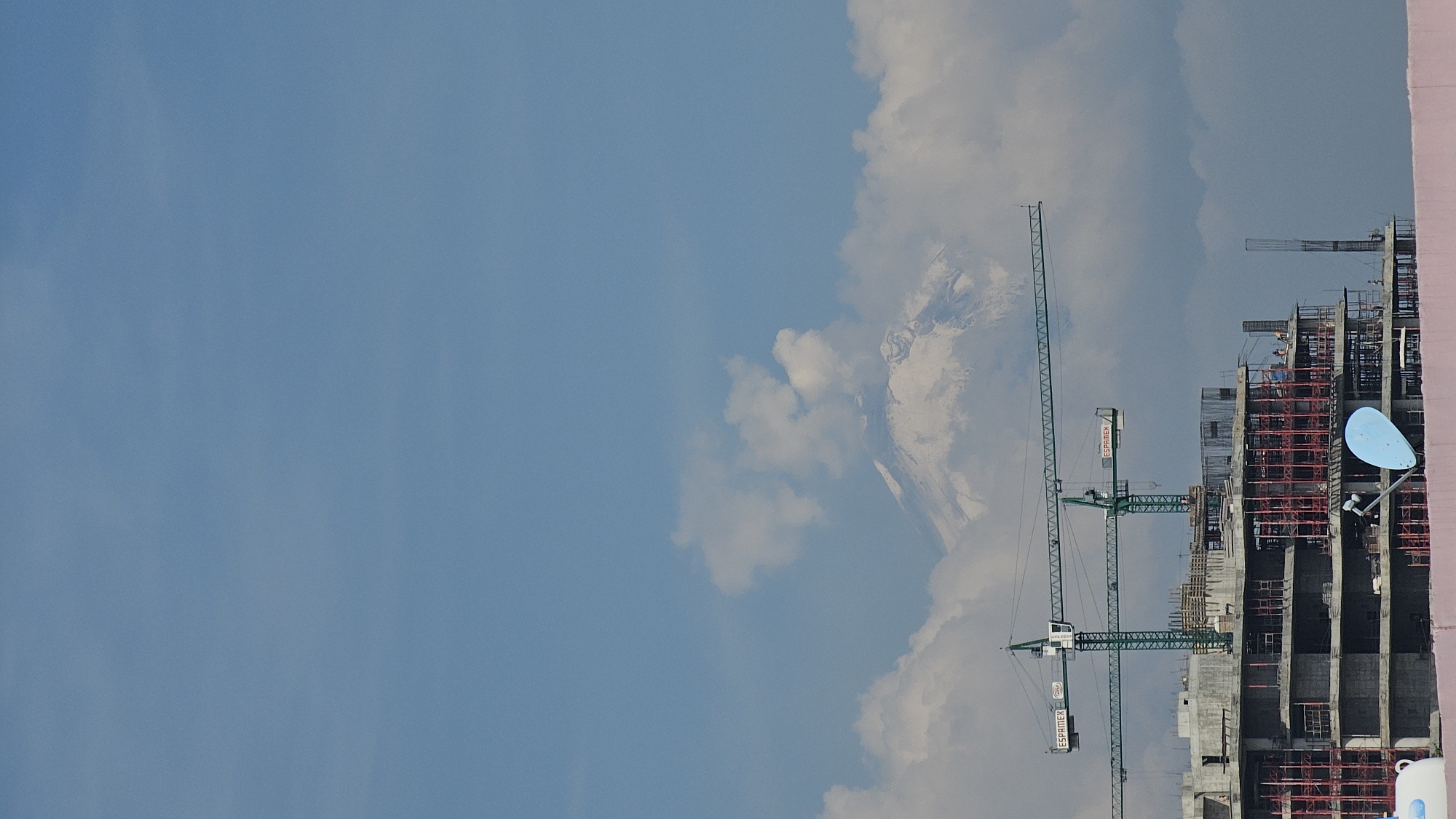The image depicts a construction site with several green cranes, likely for a high-rise building, set against a partly cloudy sky. In the background, the peak of a snow-capped mountain is visible through the clouds, suggesting a mountainous landscape near the construction site. A blue satellite dish is mounted on the side of the partially-constructed building. The scene is taken in Puebla, Mexico. The city is well-known for its proximity to the Popocatépetl volcano.