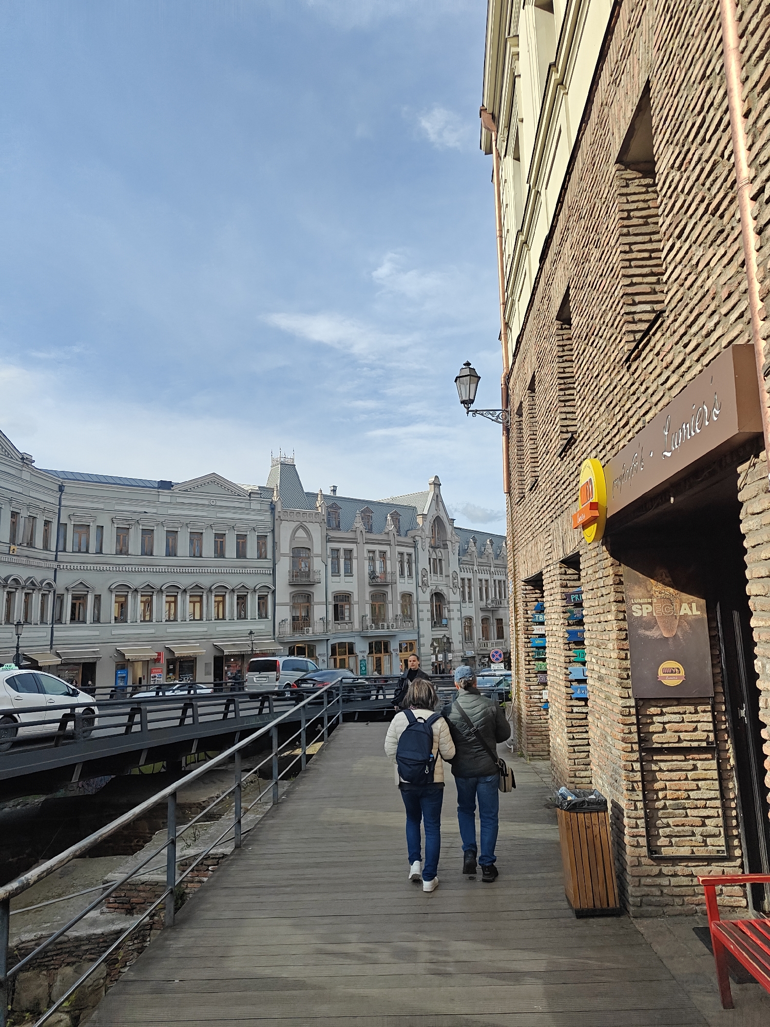 The image captures a vibrant street scene in Tbilisi, Georgia, under a bright blue sky with scattered white clouds. A wooden pedestrian walkway extends from the bottom right into the middle distance, bordered on the right by a rustic brick building. Two individuals are seen walking away from the camera on this path: one wears a light jacket and a blue backpack, while the other is dressed in a dark jacket with a shoulder bag. Further down the walkway, another person is visible.
Attached to the brick building is a classic black ornate street lamp, illuminating signs below. A wooden trash bin is positioned against the wall, and part of a red bench is visible in the lower right. To the left of the walkway, a metal railing separates it from a lower-level canal or ditch. Beyond this, a road carries several vehicles, including a white car (possibly a taxi) and a dark SUV.
Across the street, a row of grand, multi-story buildings showcases classical Georgian architecture. These structures are predominantly light grey or beige, featuring numerous windows, decorative cornices, and intricate rooflines, some with peaked towers. The scene depicts a calm daytime atmosphere of a bustling urban area.
Visible text on a large brown sign attached to the brick building reads "Lumiere's" in a cursive font, with a smaller sign beneath advertising "LUMIERE'S SPECIAL" and the Georgian word "კინო" (kino), suggesting a cinema or café. A yellow circular sign also shows "LUMI". Smaller blue signs stacked vertically on the brick wall have partial text like "PRI" and "TBI".