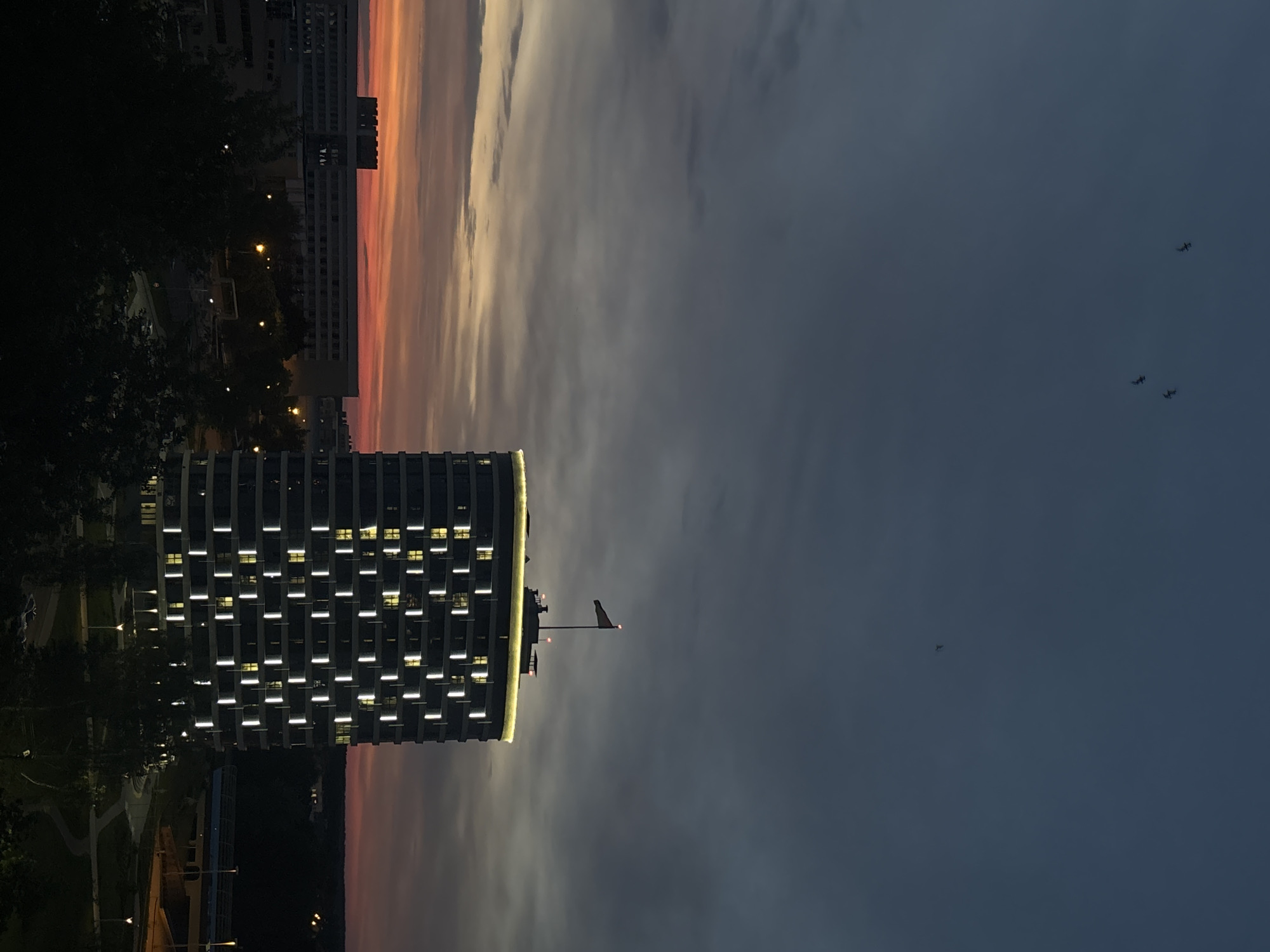 The image shows a cityscape at dusk in Minsk, Belarus. A tall, modern building with illuminated windows dominates the lower left of the frame. A flag is visible on the building's roof. In the distance, another building is partially visible with the sky appearing orange. The sky fades into a dark grey on the right side of the image. Several aircraft can be seen in the sky. A silhouetted line of trees runs along the bottom and left edges of the frame.