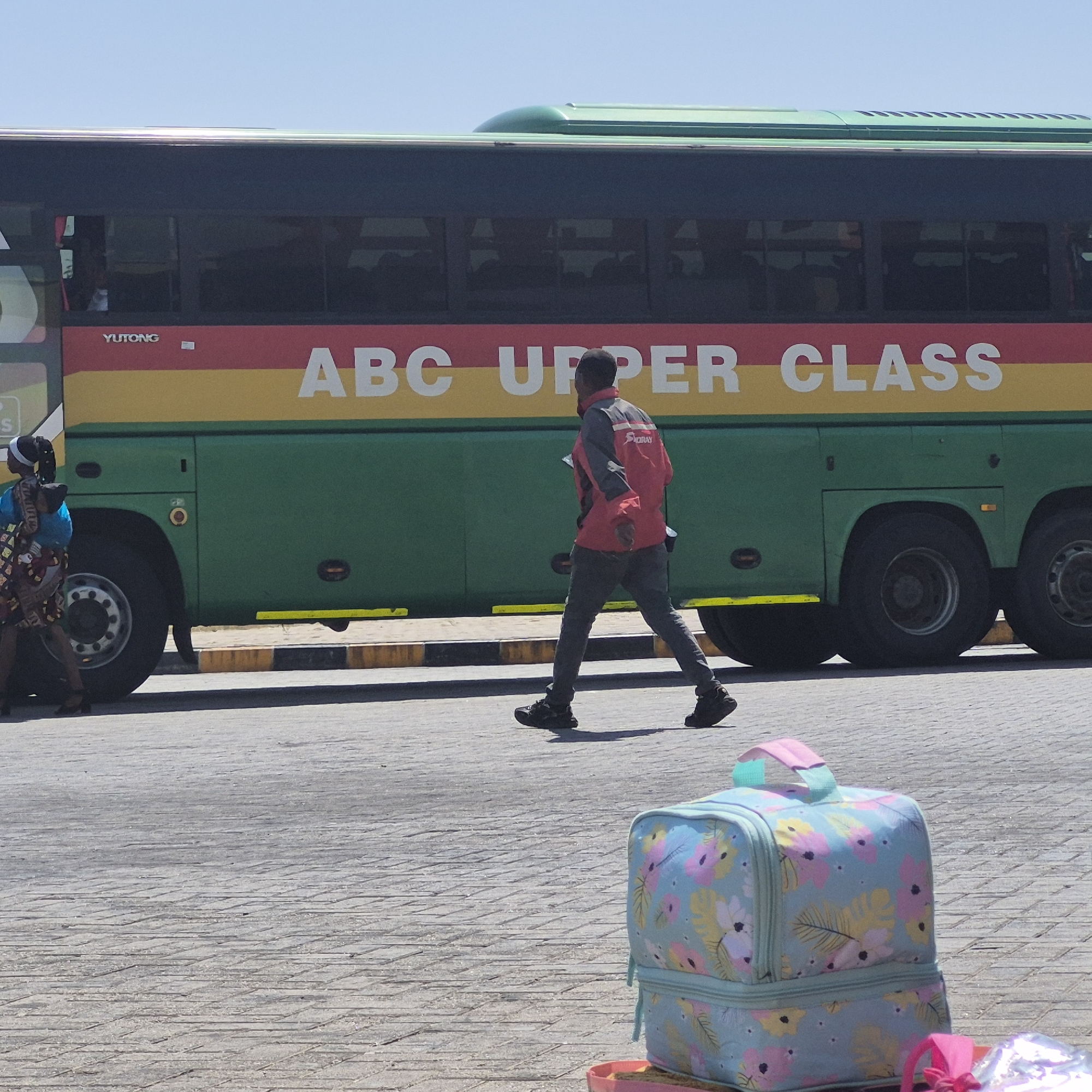 A medium-shot, eye-level, outdoor, daylight photograph captures a scene at a bus station in Dodoma, Tanzania. In the mid-ground, a green bus with a red and yellow stripe runs horizontally across its side. White lettering on the stripe spells out "ABC UPPER CLASS". A man wearing a red and gray jacket and dark pants walks across the frame from right to left, casting a shadow on the ground. To the left of the bus, a woman with dark hair pulled back and wearing a blue top and a patterned skirt walks away from the camera. In the foreground, a light blue insulated lunch bag with a floral pattern sits on the paved ground. The ground is composed of interlocking pavers. The sky above is clear and blue.