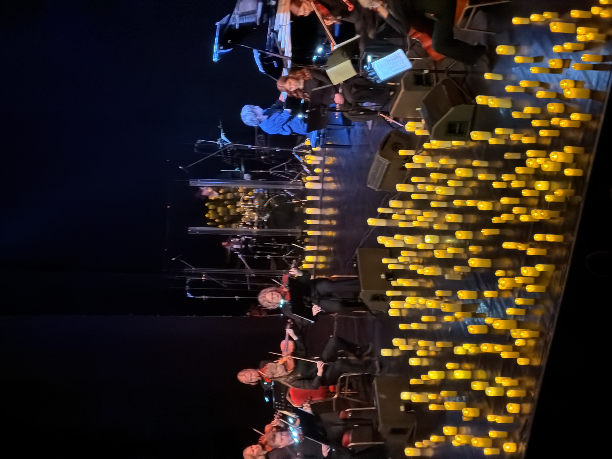 The image captures a musical performance, likely a concert, taking place on a stage. Several musicians are positioned on the stage, some playing string instruments like violins. A drum set is visible in the background, suggesting an orchestra or band performance. Notably, the front of the stage is adorned with numerous yellow, candle-like lights, creating a warm and decorative ambiance. The lighting is dim, focusing attention on the stage and the performers. The specific location within Izhevsk, Russia, is not identifiable from the image itself.