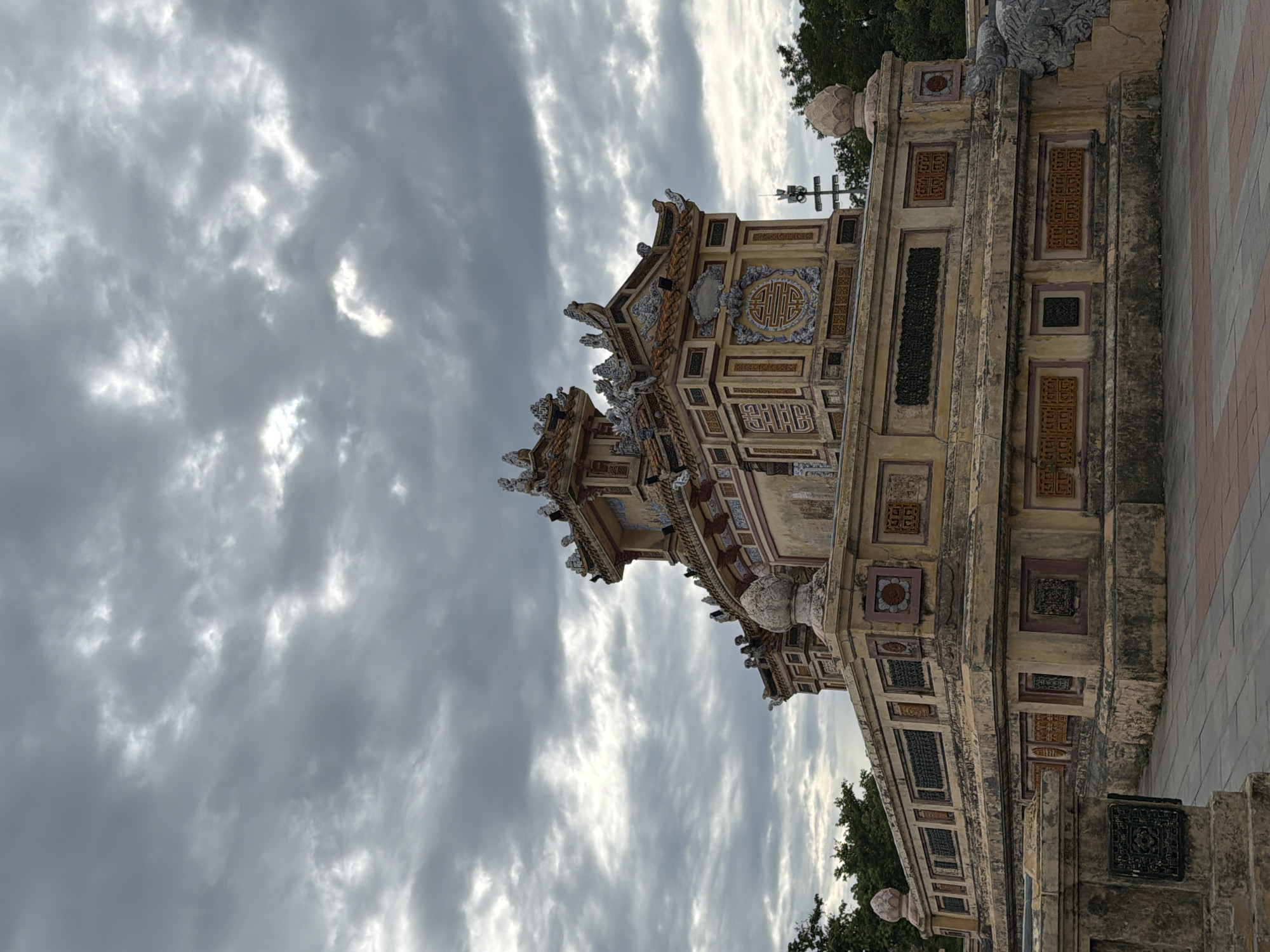 The image shows a large, ornate tomb or temple structure under a cloudy sky. The building is made of stone and is elaborately decorated with colorful tiles and intricate carvings. The structure has multiple tiers and a pagoda-like roof. The foreground shows a paved walkway leading up to the building. The background consists of trees and a tall antenna or tower. The overall impression is one of historical significance and grandeur. The tomb is located in Thuận Hóa Ward, Vietnam.