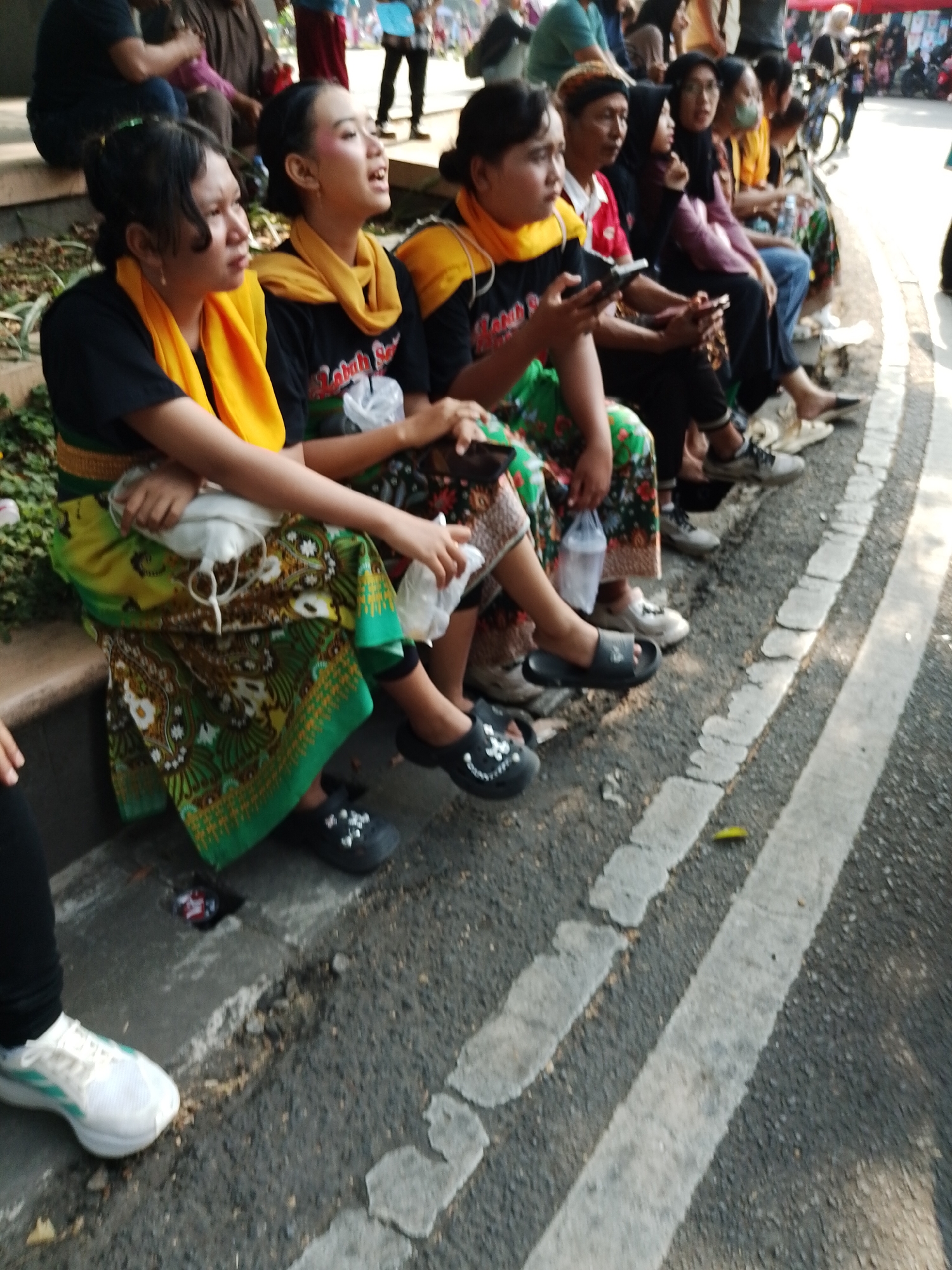 In Kecamatan Pati, Indonesia, a group of people are seated on a concrete ledge. In the foreground, two young women are prominently featured. The woman on the left wears a black t-shirt with a yellow scarf draped around her neck and shoulders, and a patterned green and gold skirt. She is looking to her left. The woman next to her, also wearing a black t-shirt and yellow scarf, is looking upwards and to her right, with her mouth open as if speaking or singing. She has makeup on her eyes and lips. Behind them, other individuals are seated, including a person in a red and white shirt and a person in a black hijab. Several people are holding mobile phones. The environment appears to be outdoors, with foliage visible behind the seated individuals. The ground is asphalt with white painted lines, suggesting it could be a roadside or a public walkway.