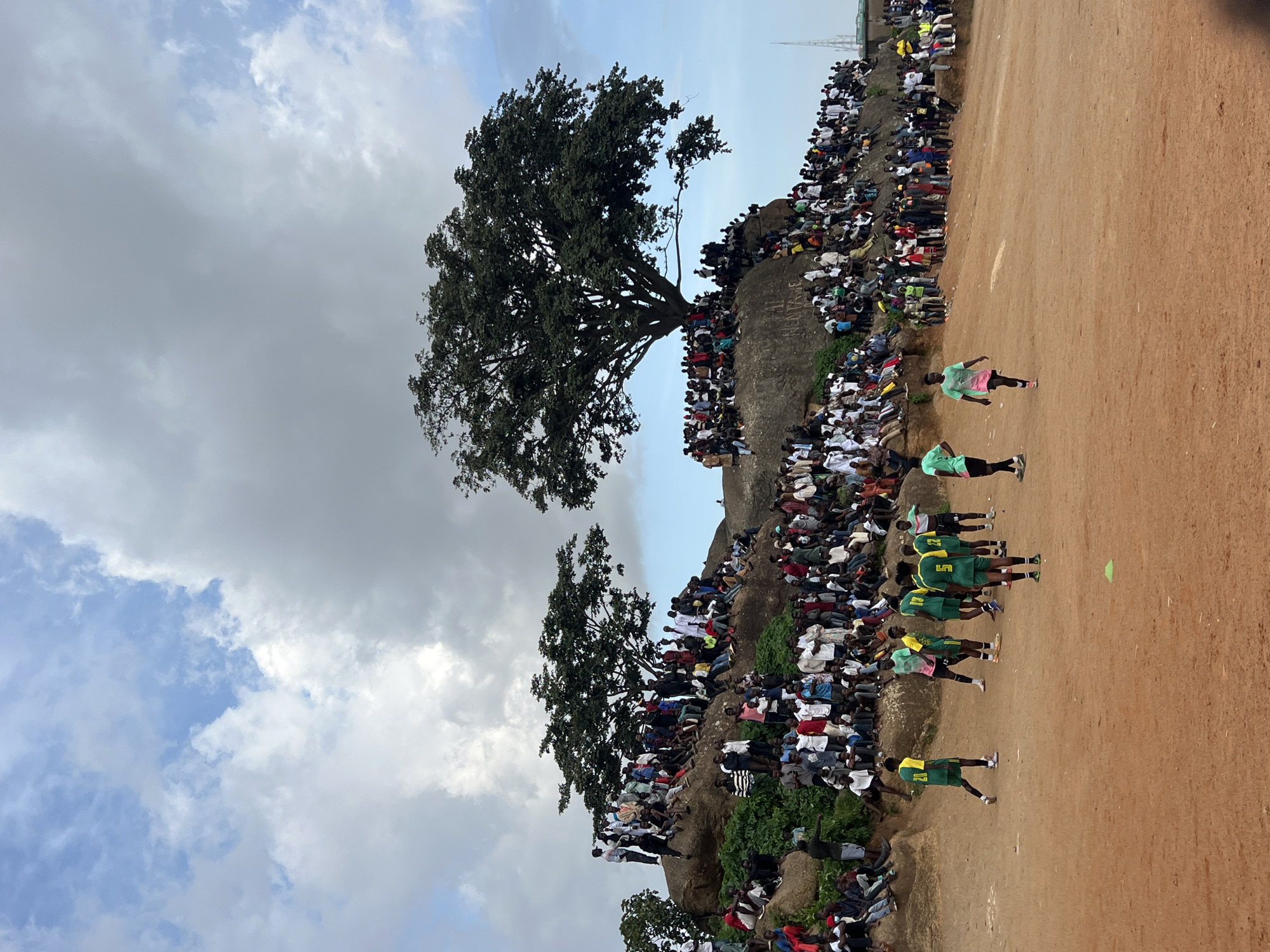 The image depicts a crowded gathering of people in the city of Jos, Nigeria, seemingly on a large rock formation overlooking a dusty field where a football match is in progress. The sky is partly cloudy, with patches of blue visible. The rock formation is densely populated with spectators sitting, standing, and leaning on the rocks. Two large trees with full canopies are situated on top of the rocks, providing some shade.
In the foreground, on the dusty field, several individuals are engaged in a football game. They are wearing athletic attire, primarily in green and yellow uniforms. Some spectators are also visible at the edge of the field. The ground is a reddish-brown earth, typical of arid or semi-arid environments. The overall scene suggests a lively community event, possibly a local football tournament or match, drawing a large audience to this unique natural amphitheater. The presence of a cellular tower in the far distance suggests the proximity of urban infrastructure.