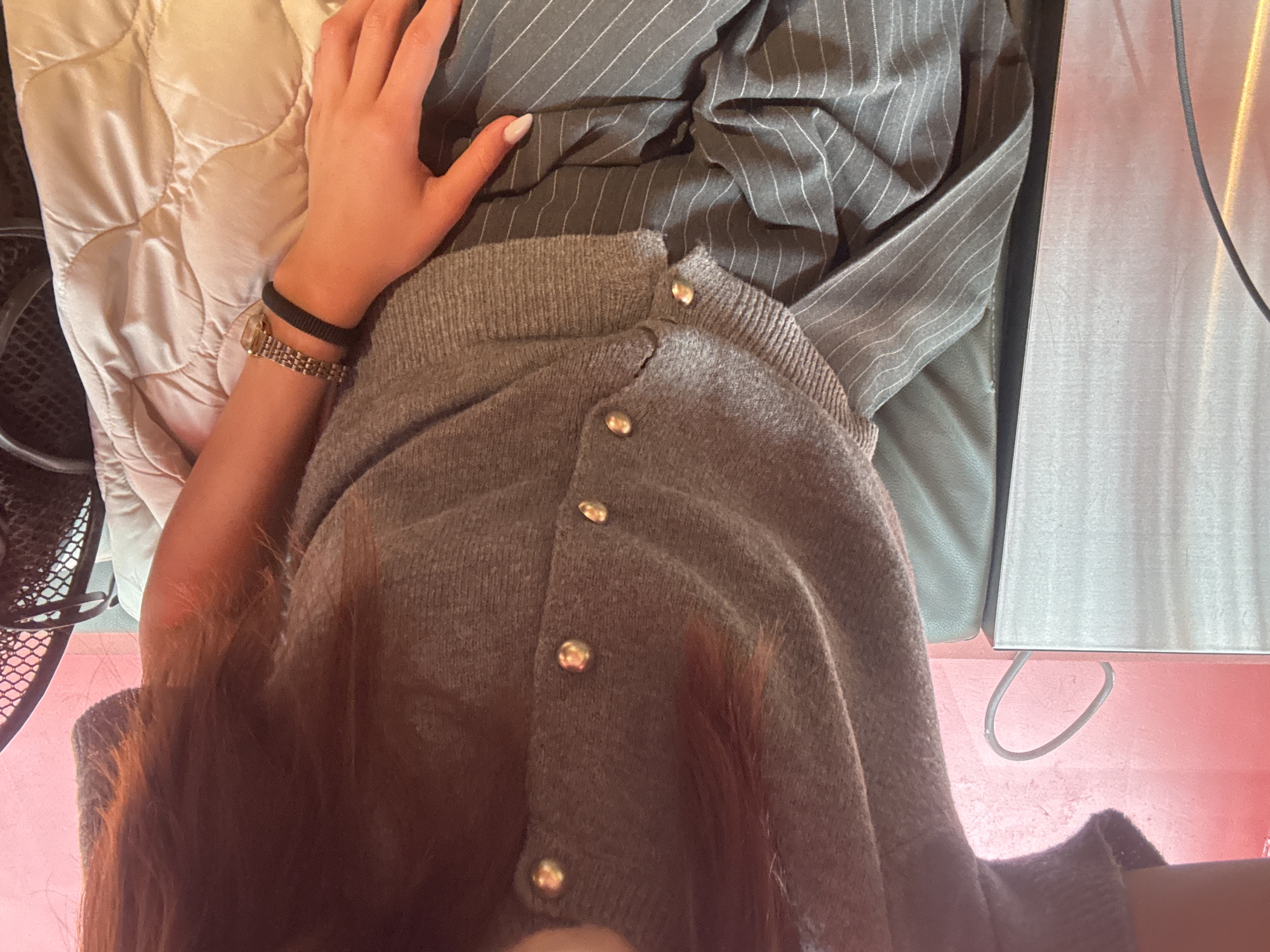 The scene presents a first-person, overhead view of a person seated in an unknown indoor location. The subject is wearing a gray cardigan with gold buttons. The person is also wearing a watch and a black wristband. A hand with painted nails is visible. The individual is seated on a cushion. A pinstripe fabric is placed on the pillow. To the right, a flat metallic object is visible on a red surface. A black fan is to the left. The city of Yerevan, Armenia, is not visually indicated in the image.