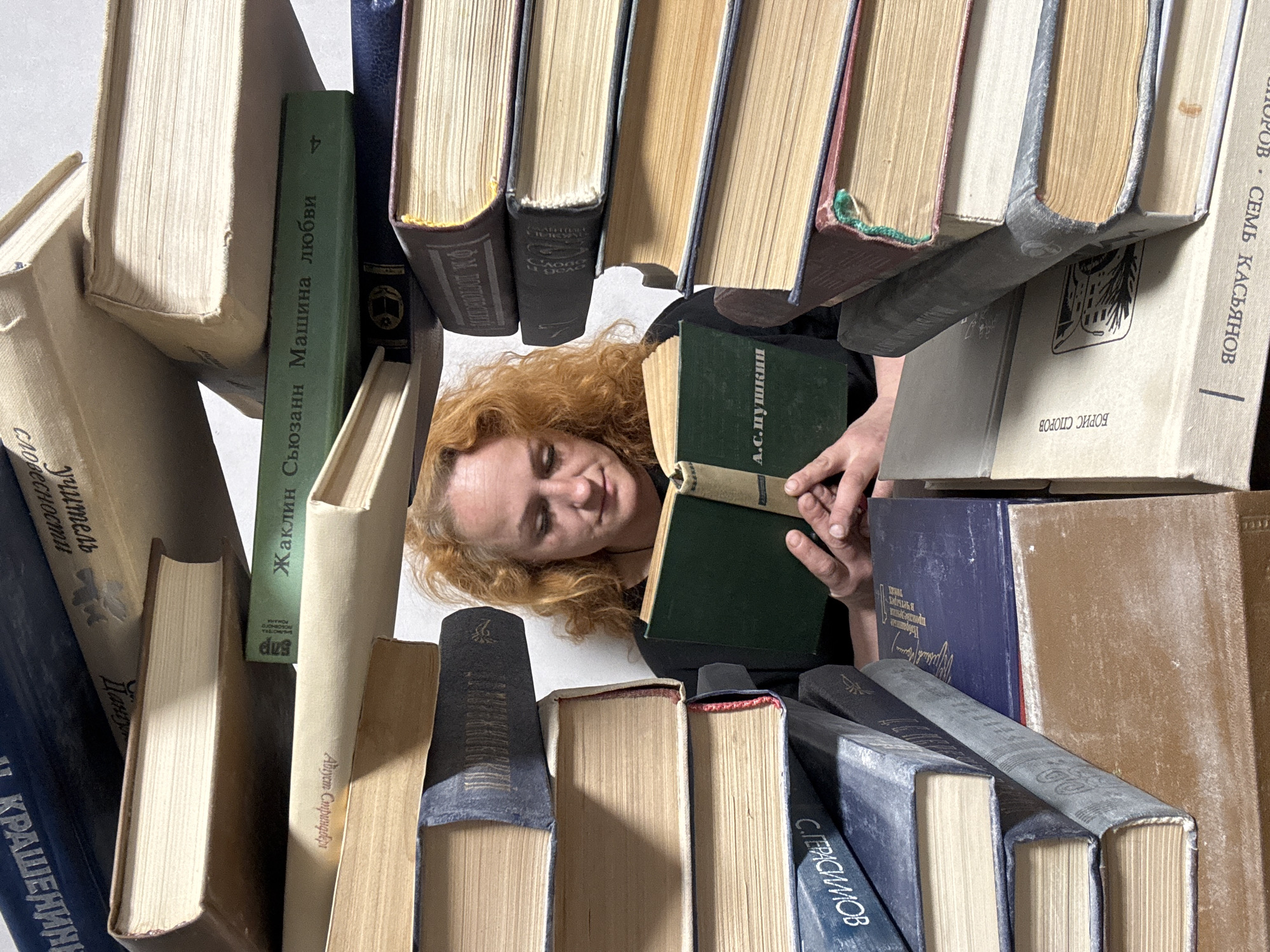 An overhead shot captures a person with curly, reddish-blonde hair immersed in a voluminous collection of books. The individual lies down, appearing calm and absorbed, holding an open, dark green book and seemingly engaged in reading. Their head is gently tilted, and a serene expression suggests enjoyment or concentration.
The scene is dominated by a multitude of books, creating a dense, cocoon-like reading environment. They are piled and arranged in various orientations—some stacked vertically, others horizontally, and many at angles—forming a chaotic yet intimate nook. The books display a range of colors, including green, blue, brown, and beige, with many showing signs of age like worn covers and yellowed pages.
Numerous book spines feature titles and authors written in Cyrillic, pointing to a collection of Russian literature or works translated into Russian. Prominently, the green book held by the person displays "А.С.Пушкин" (A.S. Pushkin). Other decipherable texts include "Жаклин Сьюзанн Машина любви" (Jacqueline Susann Machine of Love), "СЕМЬ КАСЬЯНОВ" (Seven Kasyanovs), "БОРИС СПОРОВ" (Boris Sporov), "С. ГЕРАСИМОВ" (S. Gerasimov), and "Август Стриндберг" (August Strindberg). The presence of these Cyrillic texts is consistent with the provided location context of Chaikovsky, Russia. The soft, even lighting suggests an indoor setting. The overall impression is one of profound immersion in literature.