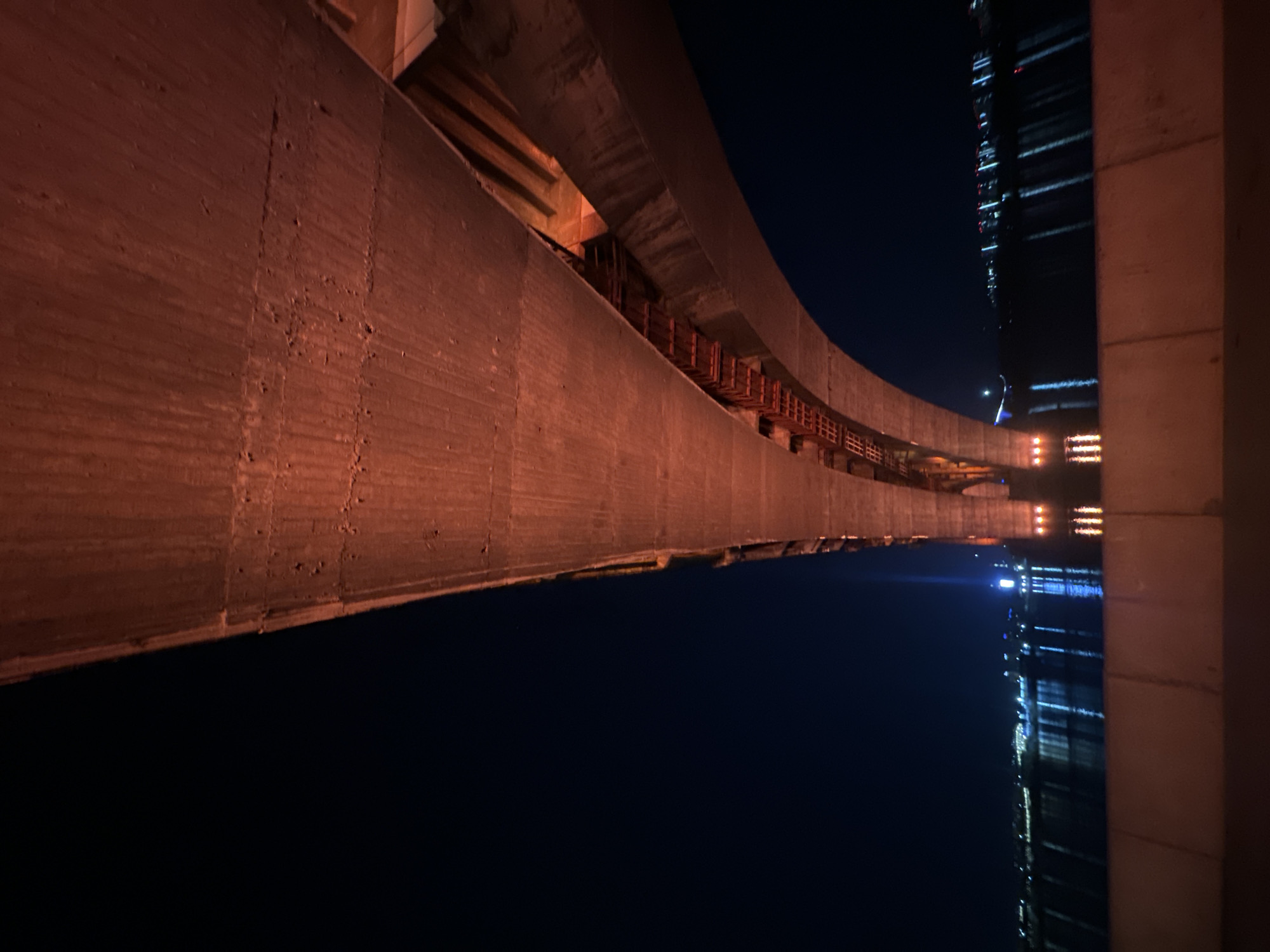 The image shows a section of the Vinogradovsky Bridge in Krasnoyarsk, Russia, at night. The bridge's concrete structure dominates the foreground, with its textured surface illuminated by an orange light source. A series of metal supports are visible beneath the bridge's span. In the background, the dark sky contrasts with a tall building reflecting lights, presumably on the Yenisei River.