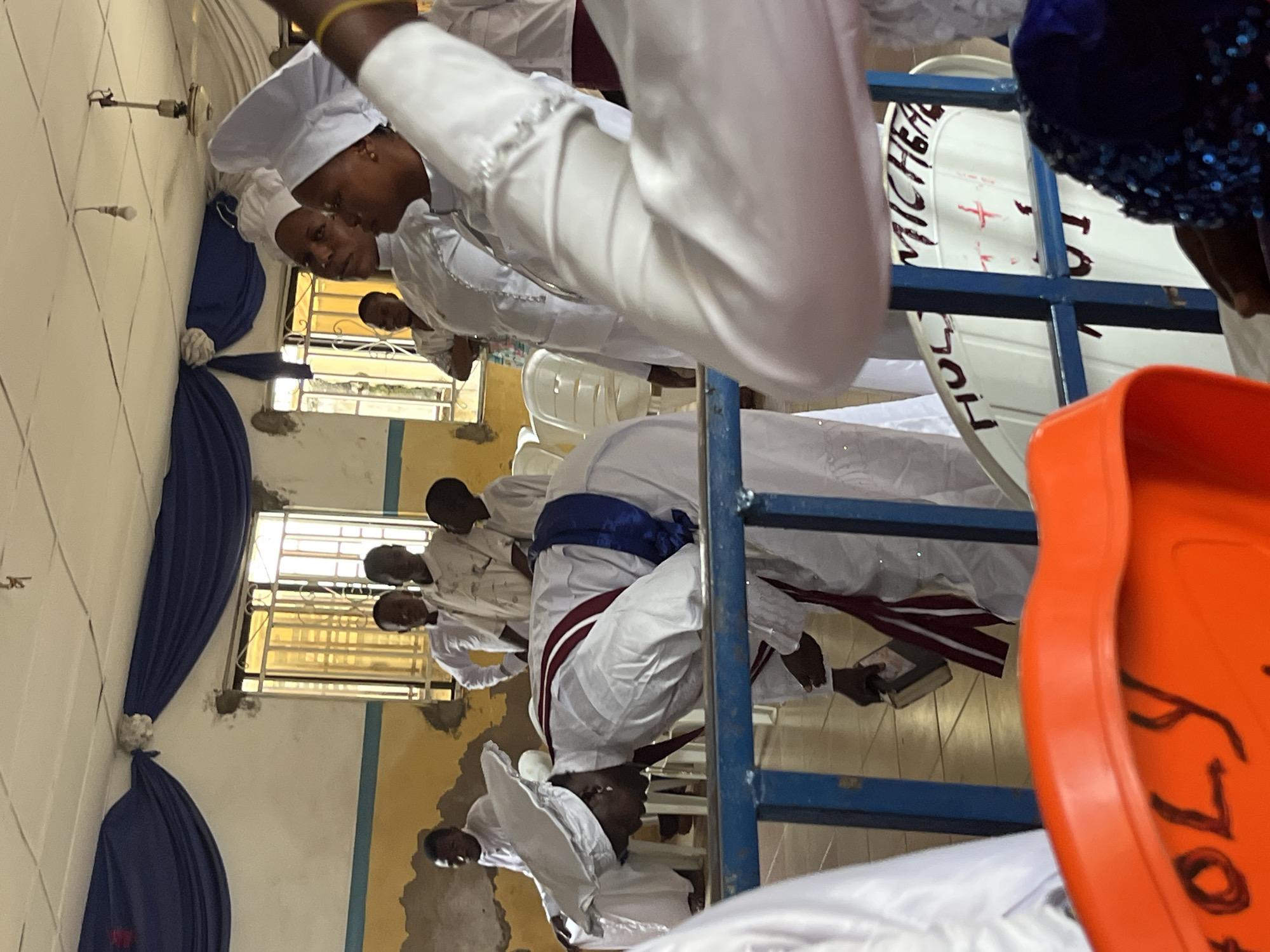 The image depicts an indoor gathering with several individuals dressed in white religious attire. In the foreground, an orange container with "HOLY" written on it is partially visible. Behind the container, a blue railing separates the viewer from the main scene. A woman in white religious attire, wearing a distinctive white head covering, is bent over, appearing to be engaged in prayer or ritual. She has a red and blue sash draped around her. Other individuals in similar white attire are standing and observing. The environment appears to be a room with yellow and white painted walls, blue drapery, and windows with metal grates. Several white plastic chairs are visible. A whiteboard or sign with text, including "HOLY MICHEAL," is mounted on a stand behind the railing. The lighting suggests an indoor setting with natural light coming from the windows. The overall scene suggests a religious service or ceremony.