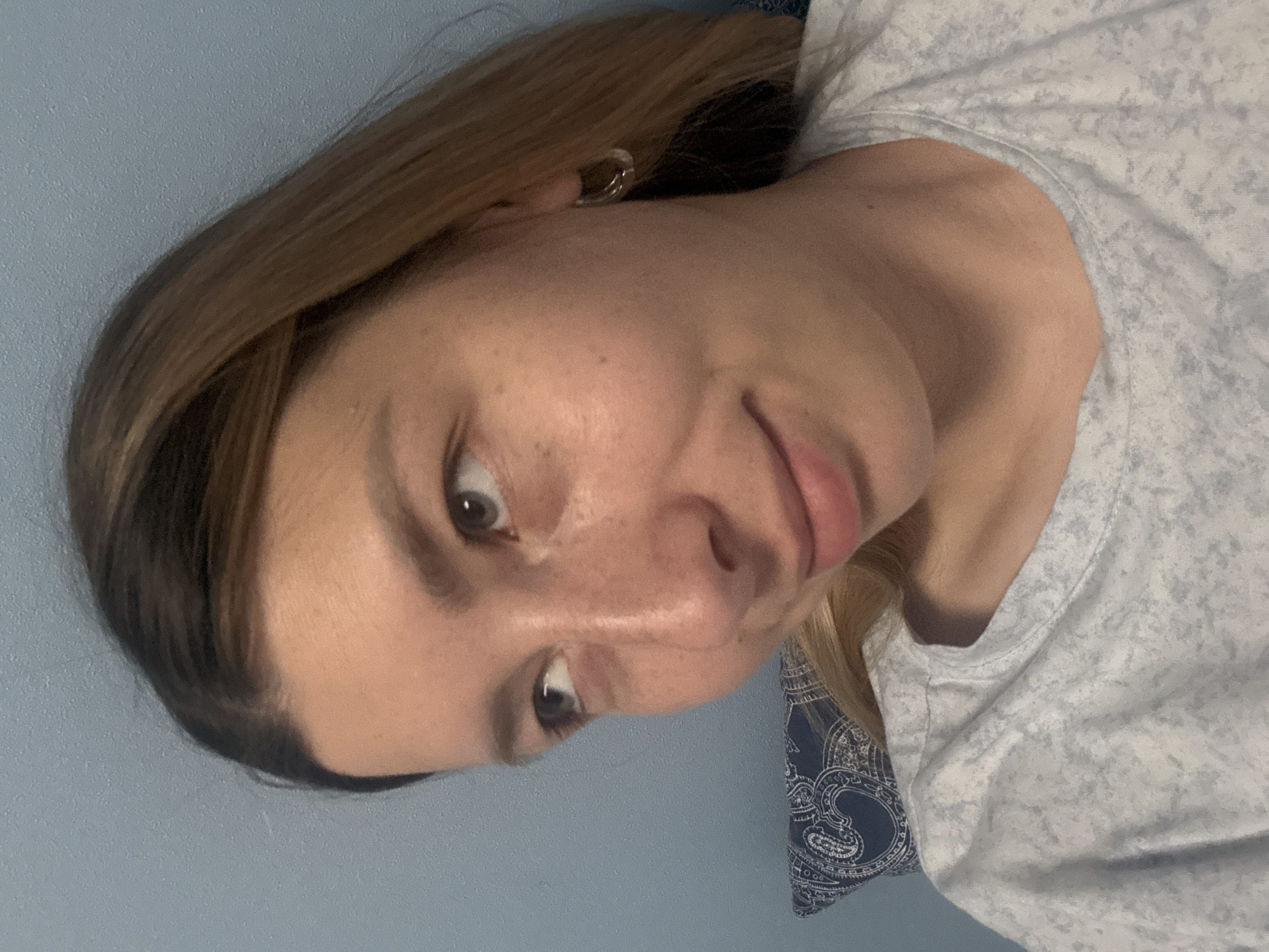A fair-skinned woman with brown hair and light eyes is facing the camera. She has a subtle smile. She is wearing a light gray t-shirt. The background is a light blue wall. There is a pillow with a paisley pattern behind her. There is no indication of Moscow, Russia in the image.