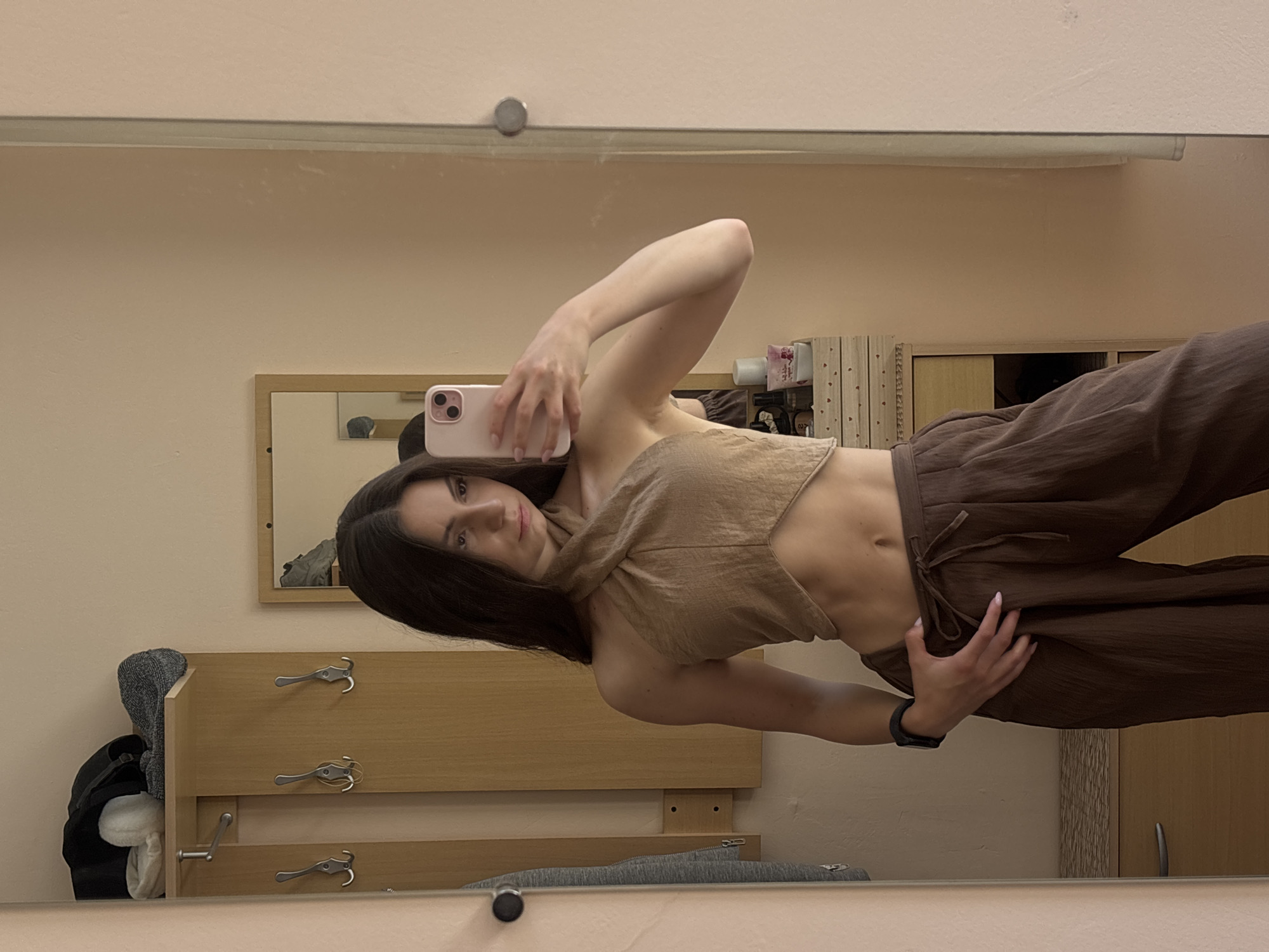 A young woman stands in front of a mirror, taking a selfie. She is wearing a brown crop top with a halter neck and wide-leg brown pants. Her hand rests on her hip. The background shows a coat rack with some clothing items and a dresser with a mirror reflecting part of the room. The scene is indoors, likely in a residential setting, with neutral-colored walls. There is no identifiable information suggesting the location is Varna, Bulgaria.