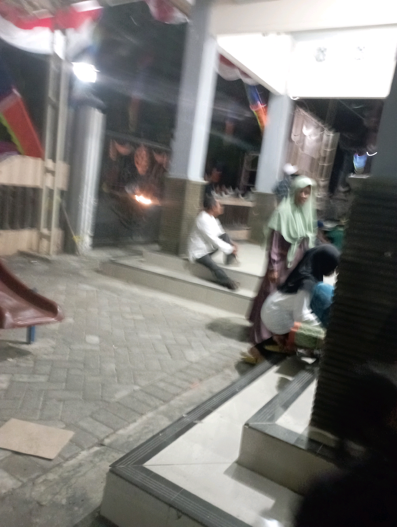 The image depicts an outdoor scene at dusk, possibly in a public area or community space in Ketapang Lor, Indonesia. In the foreground, a patterned tiled walkway leads to a set of stairs. To the left, a red slide is partially visible.
Several individuals are present. In the mid-ground, a person wearing a light-colored shirt and dark pants sits on the stairs, facing left. Next to them, a person in a light green hijab and a long, patterned dress stands. In front of the person in the hijab, another individual, wearing a white shirt and dark pants, is crouched down, facing away from the camera. A person in a black hijab is also partially visible, crouching near the second individual.
In the background, a building structure with pillars is visible, decorated with red and white flags and streamers. Lights are on within the building, casting some illumination. A glowing orange light source, possibly a fire or a bright lamp, is visible in the dark recesses of the background structure. The ground is paved with interlocking rectangular stones. The overall lighting suggests it is either early evening or a dimly lit area.