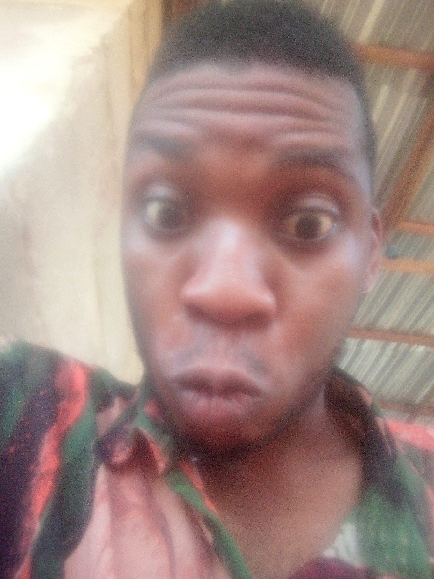 A close-up selfie shows a Black man with dark hair and brown eyes, puckering his lips. He is wearing a shirt with green, red, and pink patterns. The background consists of a wall and a corrugated metal roof. The location is Unknown, Unknown.