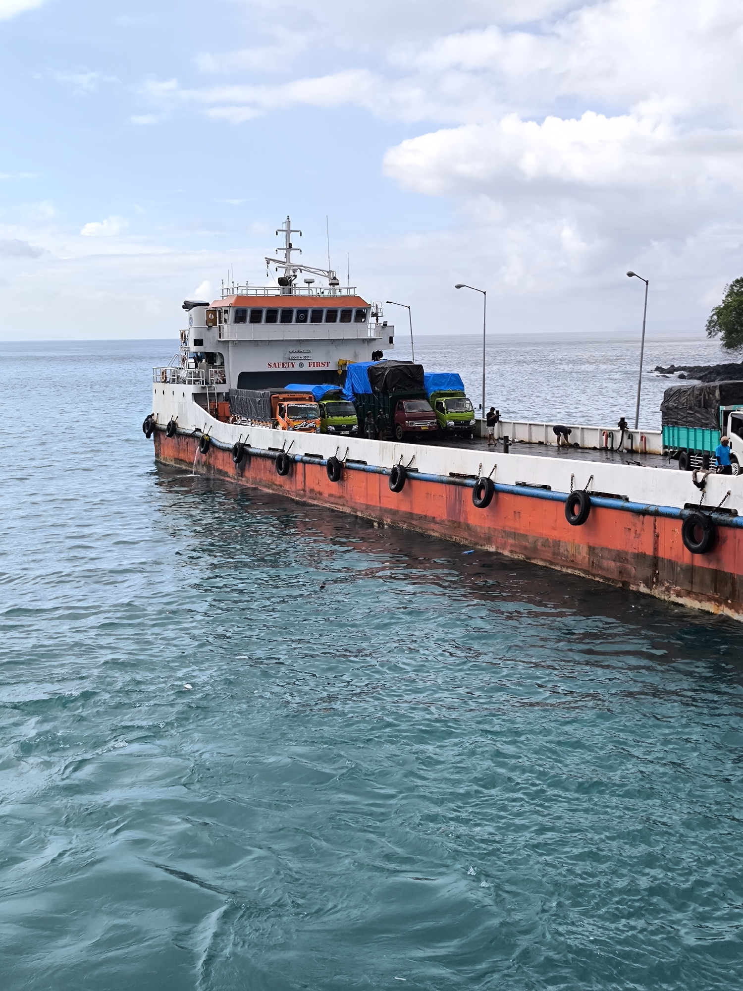 A ferry is docked on a flat, calm sea, with trucks lined up on its deck. The ferry has a two-story structure with "SAFETY FIRST" written on it. Black tires hang along the ferry's side as buffers. Streetlights are spaced along the deck. The sky is partly cloudy, and land with a few trees is visible in the distance to the right. The image appears to be taken from a similar height level on a dock or another vessel. The location is Unknown, Unknown.