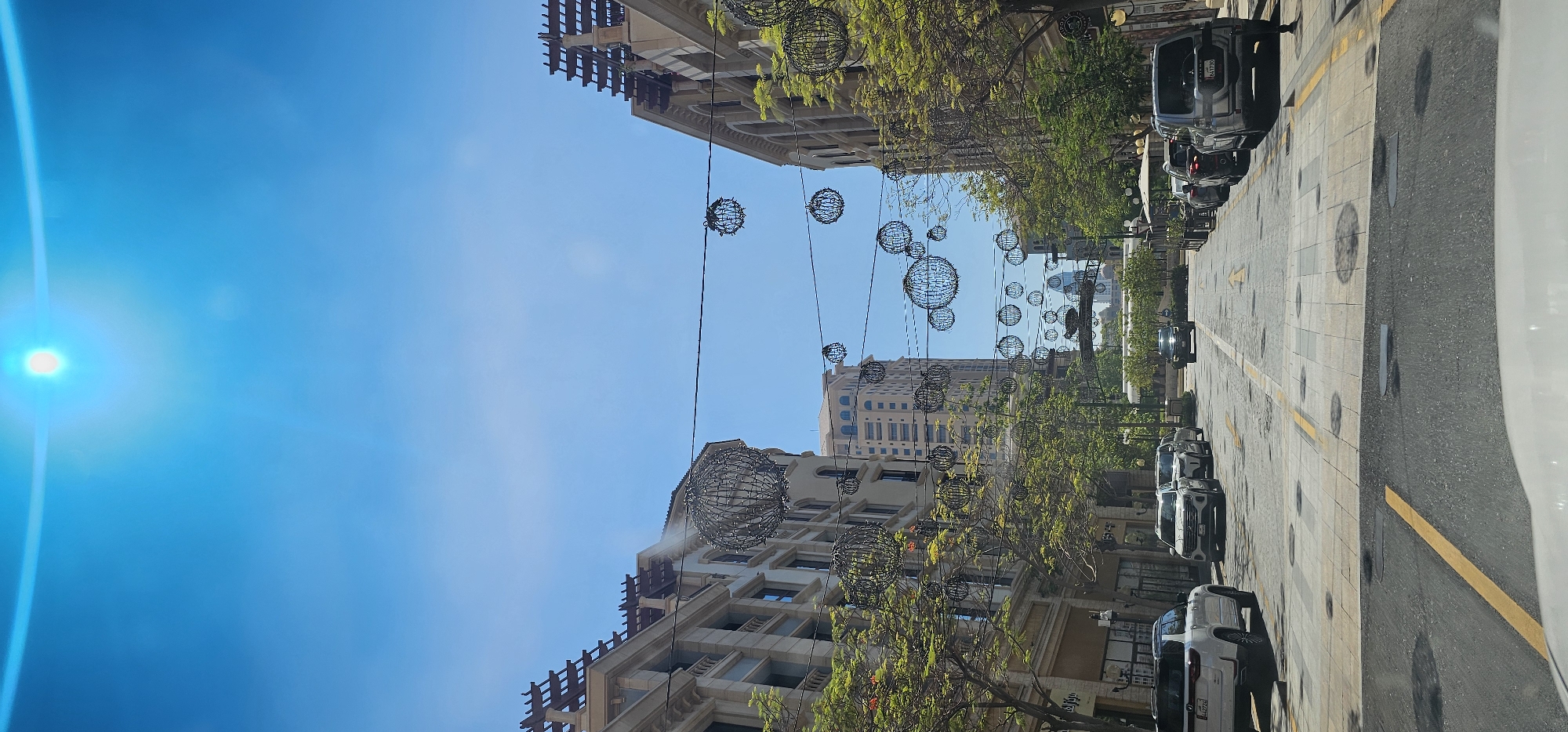 The image depicts a sunny day in an urban setting, likely Doha, Qatar, as indicated by the architecture and the presence of what appear to be decorative spherical lights hanging from wires across a street. The sky is a clear, bright blue with a noticeable lens flare from the sun on the left side of the image.
The scene is a street lined with modern buildings, some with intricate architectural details. Trees with green foliage are planted along the sidewalks, casting dappled shadows. On the right side of the street, several cars are parked. The street itself has a pedestrian walkway made of patterned tiles, and an asphalt road marked with yellow lines. The spherical lights, some larger and some smaller, are suspended at various heights, creating an ornate canopy over the street.
The overall impression is one of a vibrant, well-maintained urban environment on a beautiful day. The light suggests it is daytime, likely late morning or early afternoon given the intensity of the sun. There is no visible text in the image, and no people are clearly discernible. The scene conveys a sense of pleasant weather and an aesthetically pleasing streetscape, possibly a commercial or residential area designed for walking and leisure.