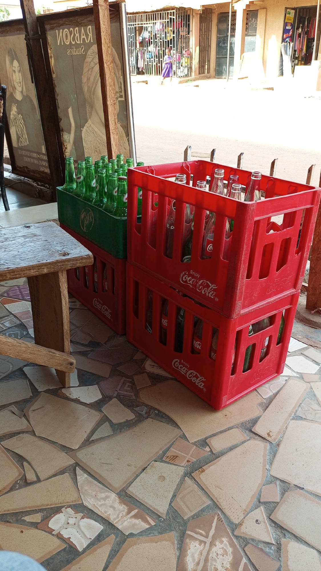 The image presents an outdoor commercial scene in Sabon Tasha, Nigeria, under bright daylight. In the foreground, a wooden table or bench is partially visible on the left. The ground is paved with an irregular mosaic pattern of light-colored tiles.
The main subjects in the midground are stacks of empty beverage crates. On the left, two green plastic crates, one displaying "Coca-Cola" branding despite containing green glass bottles, are stacked. Adjacent to them on the right are two red plastic crates, clearly branded "Enjoy Coca-Cola" and featuring the "Coca-Cola" logo, filled with empty clear glass bottles.
In the background, two large, framed posters lean against a wall. The text on these posters is mirrored, revealing "RABSON Studios" ("ИОГНАЯ zoibut2") and other details like "2HO12-AIDEO COFENICE", "ROMI HARVA", and "LAMENAGER". Beyond this area, a street scene is visible with buildings that appear to be shops or market stalls. One shop features a metal grate pulled across its entrance, with clothes visible behind it. A small child in a purple dress is seen walking in the distance on the sunlit street. The scene depicts a common commercial setup where bottled beverages are sold and empty bottles are collected.