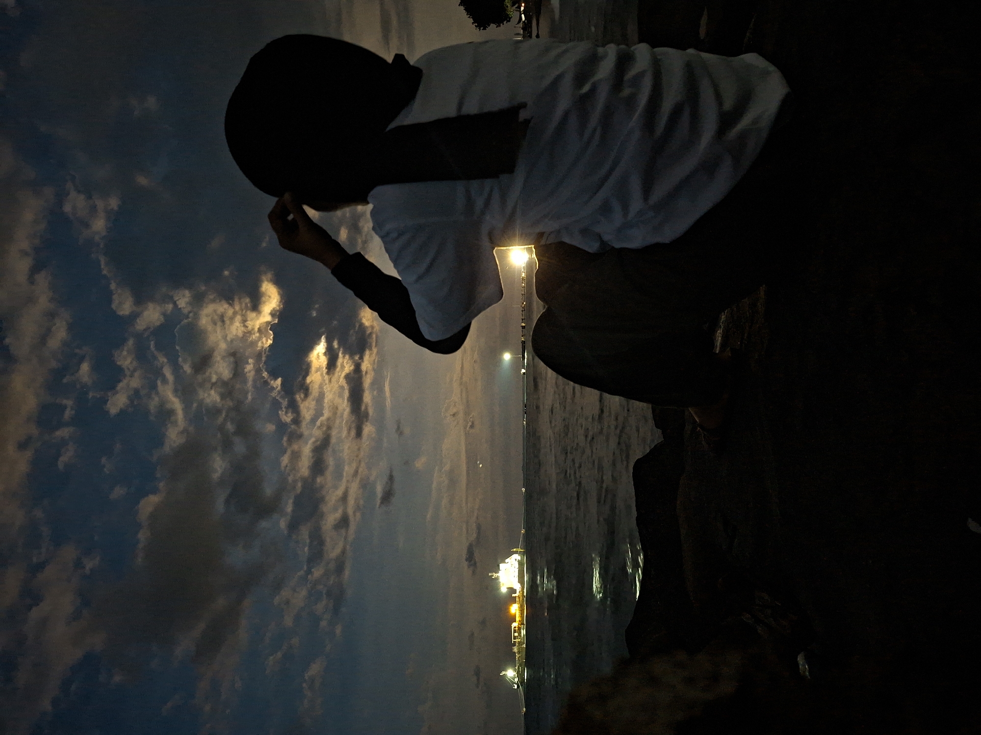 A person is crouching on a dark surface, possibly rocks, near the water at night in Lhokseumawe, Indonesia. The sky is visible, with a mix of clouds illuminated by the moon or other light sources. The person is wearing a white t-shirt and a dark head covering. In the distance, there is a large ship docked at a refinery or port, with lights reflecting on the water. The light source is causing a glow in the sky above the ship. The person seems to be looking out at the water or the sky.