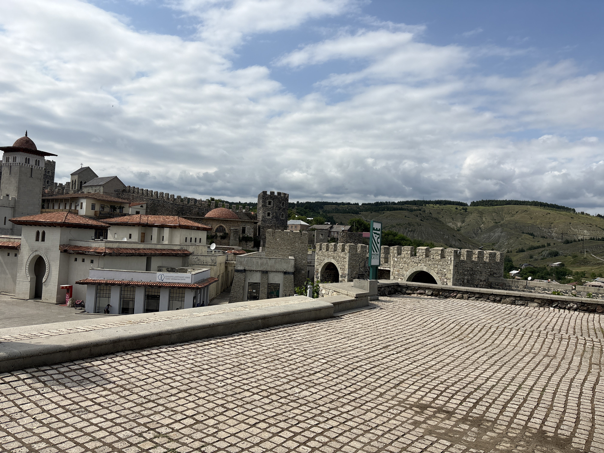 The image depicts the Akhaltsikhe Castle in Akhaltsikhe, Georgia, on a partially cloudy day. The foreground is dominated by a cobblestone plaza. In the midground, a complex of stone buildings with red tile roofs is visible, including a large tower with a domed roof and arched windows, and a fortified wall with crenellations. To the right of this, another section of the castle features stone archways and battlements. A green sign with white lettering reading "Rabati Castle" is also visible. In the background, rolling, green hills stretch towards the horizon under a sky filled with scattered white clouds. A few people are visible in the distance, near the buildings.