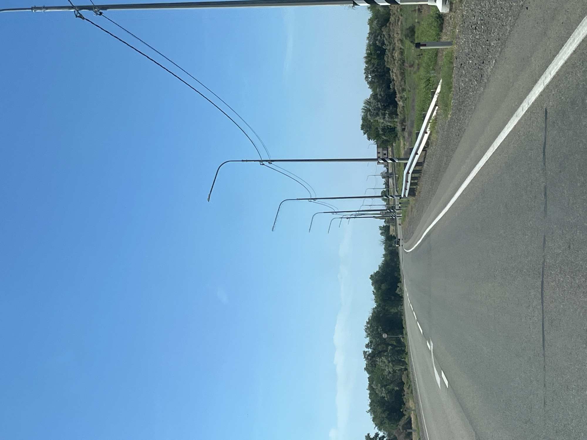 The image shows a two-lane asphalt road winding through a rural landscape under a clear blue sky. Streetlights line the right side of the road, their poles interspersed with power lines. To the left of the road, there is a dense line of green trees, and to the right, a grassy embankment with a metal guardrail. A single black car is visible in the distance on the right lane, heading away from the viewer. The road surface appears to be in good condition with white lane markings. In the background, more trees and some low buildings are visible. This scene is consistent with a road in the vicinity of Bakhtemir, Russia, likely an area with open terrain and scattered vegetation.