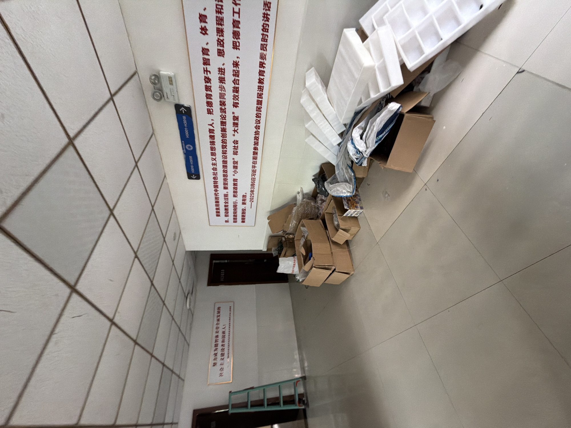 The image depicts an interior hallway with white tiled floors and a drop ceiling. On the left, a green ladder is leaning against a wall. A white sign with Chinese characters is mounted on the wall to the left of the ladder. Further down the hallway, there are two doors, one labeled "H3033". The right side of the hallway is cluttered with various cardboard boxes, some open and some sealed, along with white foam packaging materials. A long white sign with Chinese text is prominently displayed on the wall above the boxes. Above this sign, a blue directional sign with text in both English and Chinese is mounted. The environment appears to be a school or institutional building, possibly undergoing renovation or clearing out. The lighting is overhead fluorescent lighting.