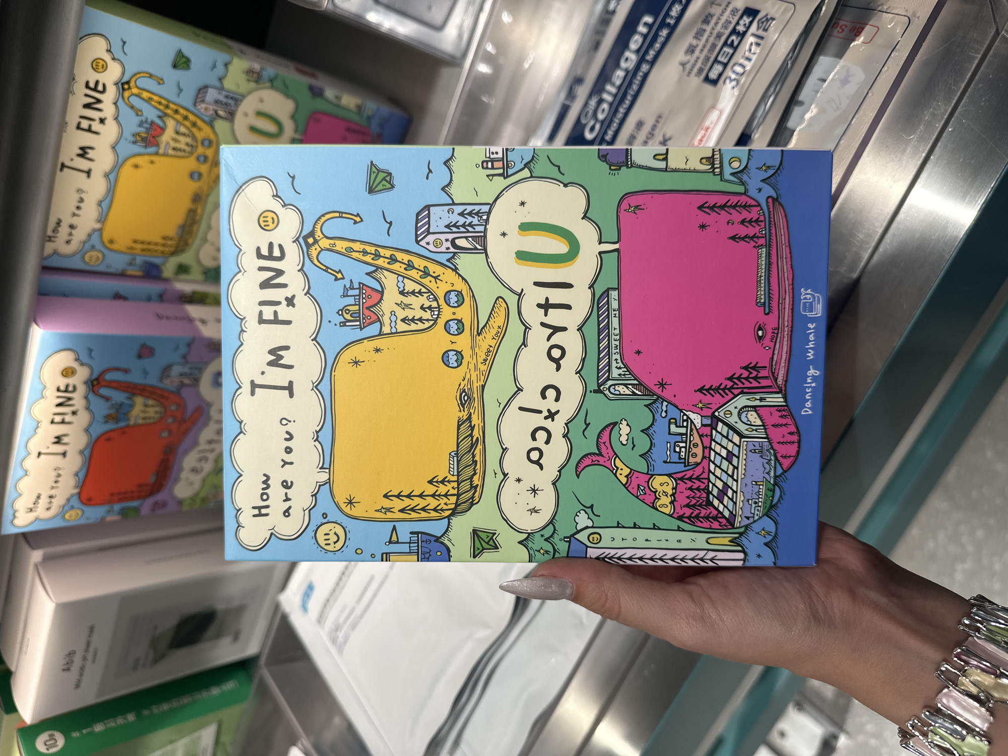The image shows a hand holding a colorful book with a drawing of two whales and text that reads "How are you? I'm FINE." The book is placed on a shelf in a store. In the background, there are other products on display, including skincare items. The setting appears to be a retail environment, possibly in Futian District, China, given the presence of some Chinese text on the products in the background. The hand is adorned with a bracelet.
