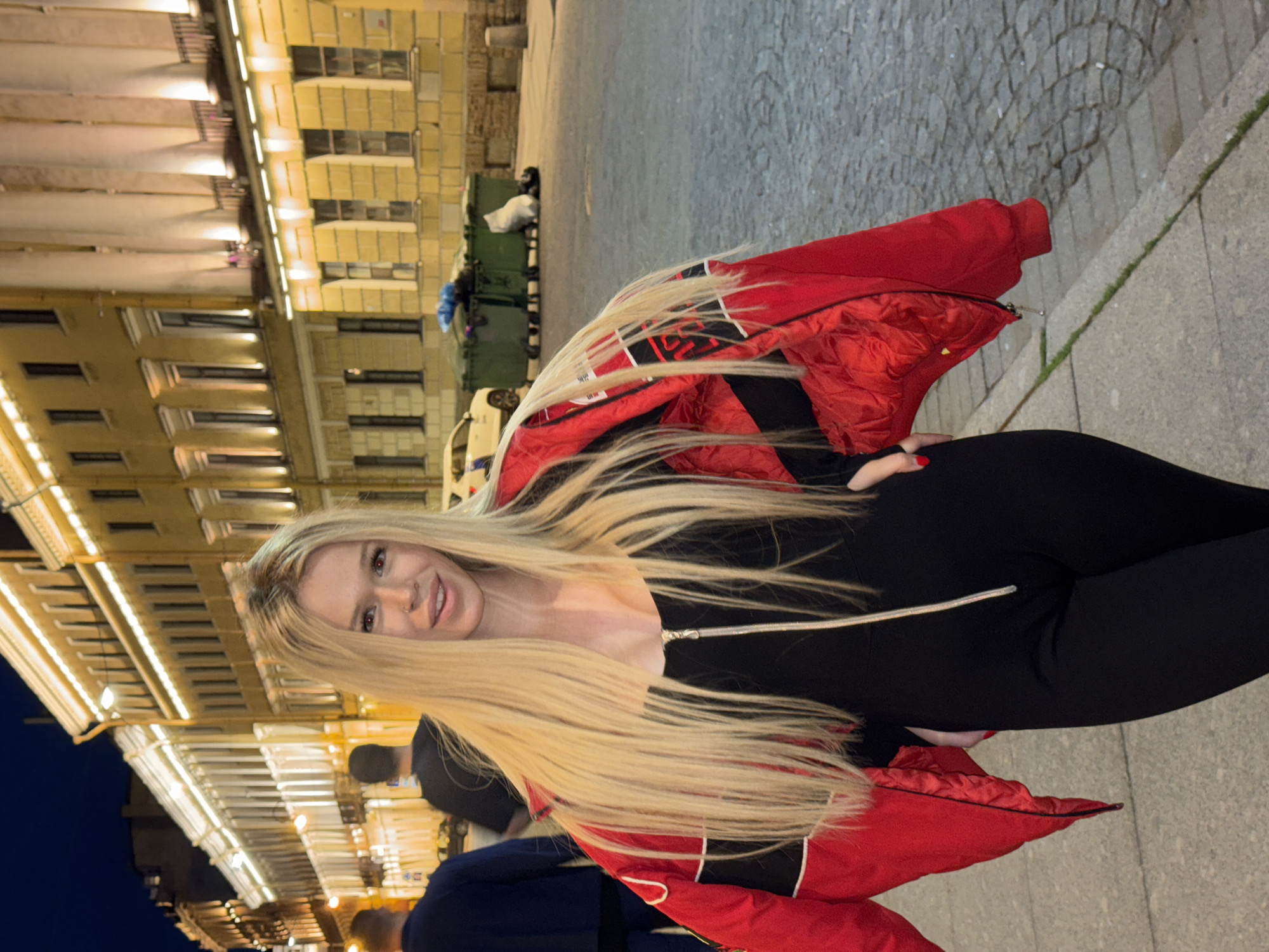 The media file shows a young woman with long blonde hair, smiling at the camera. She is wearing a black outfit and a red jacket with black and white accents. The background is a street scene at night, with buildings illuminated by warm lights. Paved sidewalks and streetlights are visible. There are some large green dumpsters in the background, and a car parked on the street. The overall impression is of a casual evening outing in an urban environment. The lighting suggests it is nighttime, and the weather appears to be clear. The architecture of the buildings hints at a European setting, and the specific mention of Saint Petersburg, Russia provides geographical context. The woman's expression is happy and engaging, suggesting a positive mood.