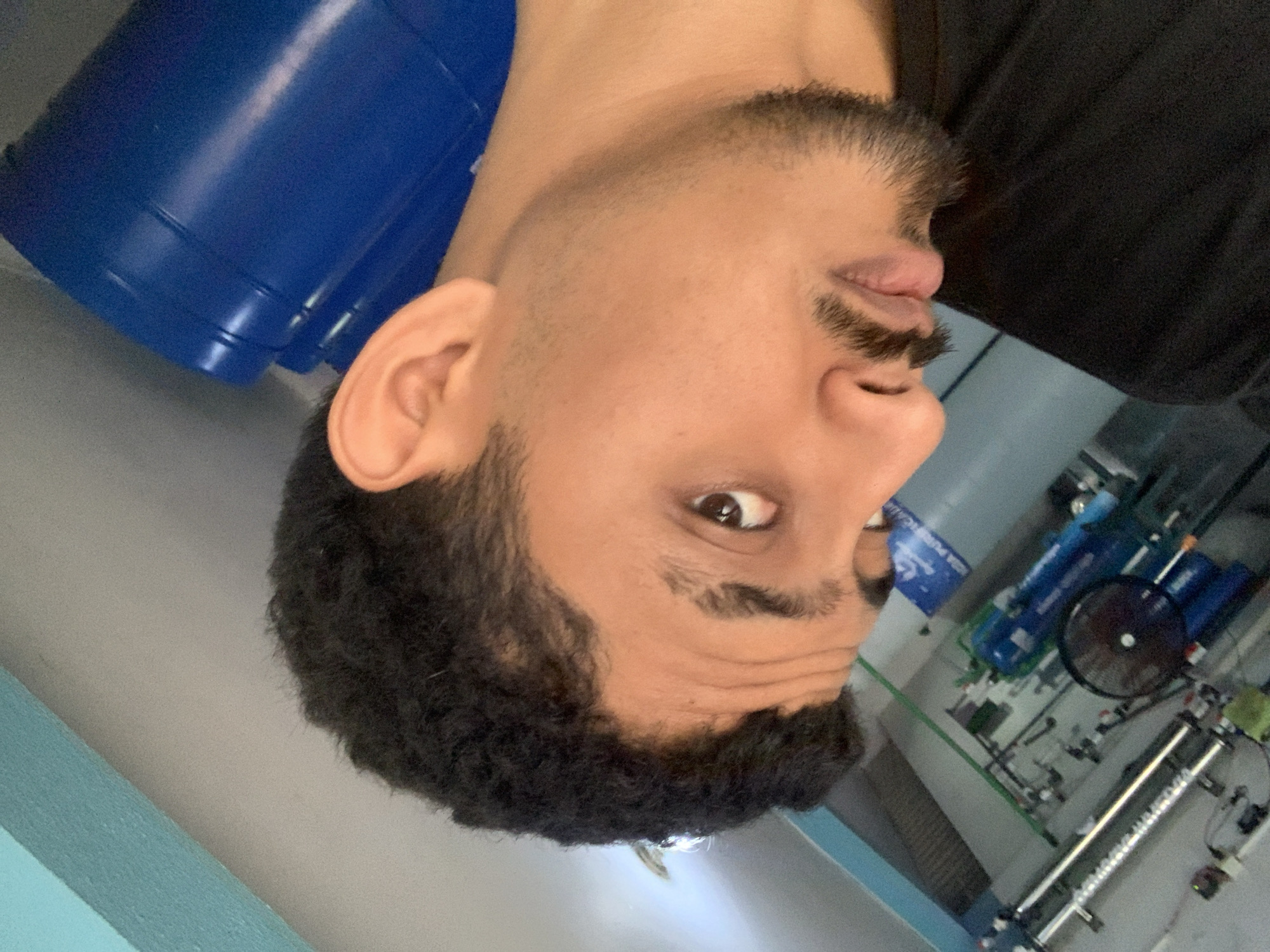 An upside-down, close-up portrait features a young man with medium skin tone, short curly dark hair, and a trimmed beard and mustache. His dark eyes are open and look directly towards the camera. Slight wrinkles are visible on his forehead. He is wearing a dark-colored shirt, with only a portion visible.
The background suggests an industrial or utility room, likely a water purification plant. To the left side of the frame (which would be the top in a right-side-up orientation), large blue plastic containers or barrels are stacked. On the right side of the frame (the bottom in an upright orientation), there is an intricate system of pipes, gauges, and what appears to be water filtration or treatment machinery. A black industrial fan is mounted amidst the equipment, and a long, metallic cylindrical unit, possibly a UV sterilizer or filter, is prominent. A blue sign on the wall in the background shows partially visible text, including "ADA PURIFICADA," strongly indicating a purified water context. The walls are light grey, with accents of green and blue structures. The indoor setting implies a daytime environment in Tacarigua, Venezuela.