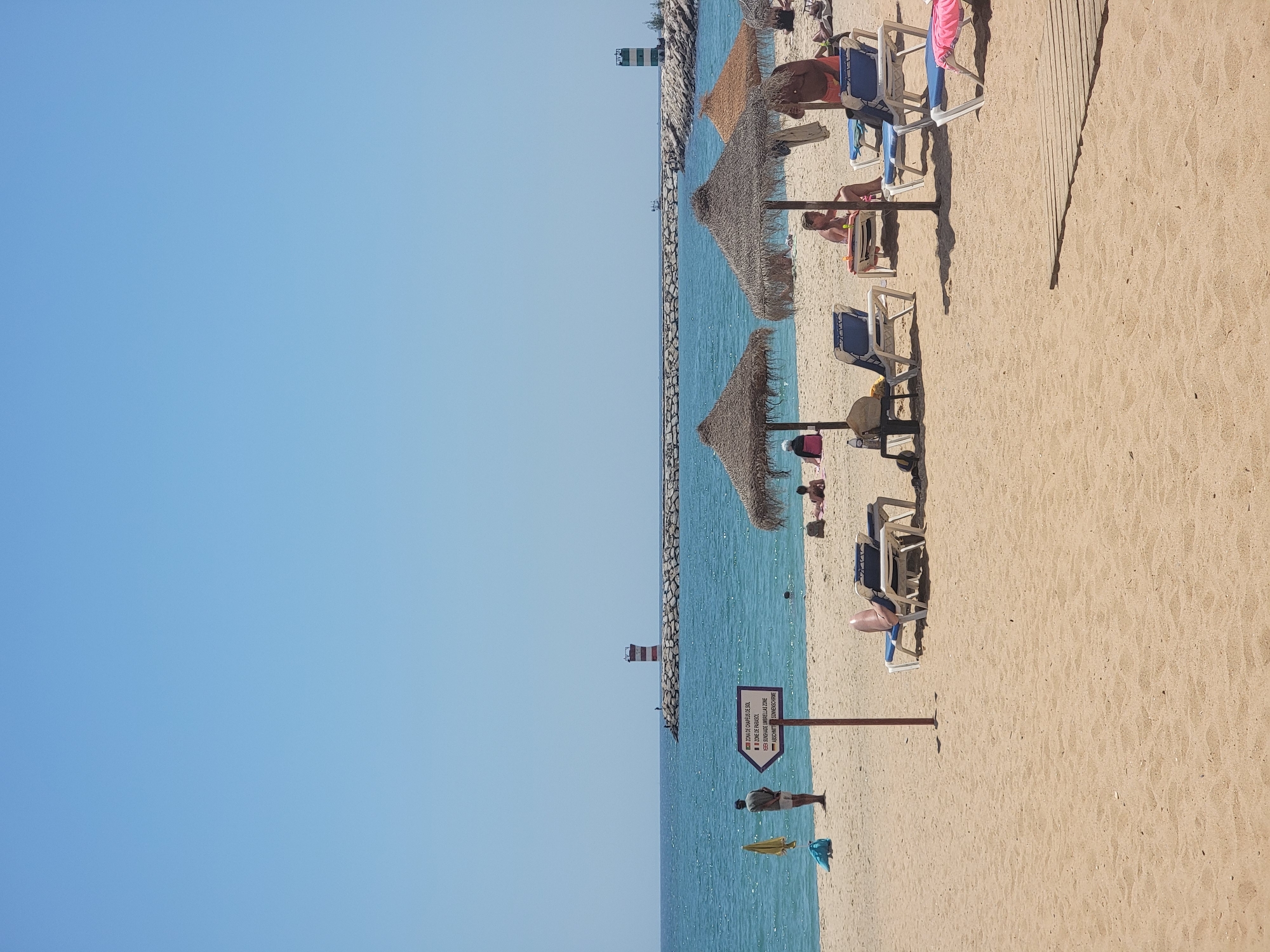 The image shows a sandy beach with the ocean in the background in Quarteira, Portugal. The sky is clear and blue. In the distance, a stone jetty extends into the sea, with two lighthouses on it. One lighthouse is red and white striped, and the other is green and white striped.
On the beach, several umbrellas and lounge chairs are set up. Some people are relaxing on the chairs, while others are standing or walking. There is a signpost with an arrow pointing towards a designated umbrella zone. The sand appears to be clean and well-maintained. The water is calm and appears to be a clear blue color. The overall atmosphere is one of a relaxed and sunny beach day.