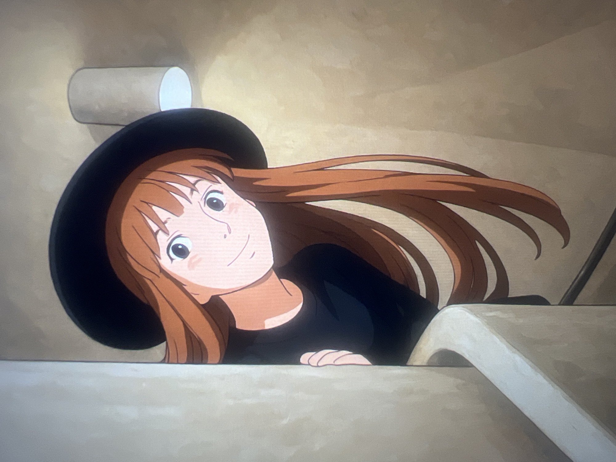 The image displays an animated young woman with long, light brown hair and fair skin, wearing a black wide-brimmed hat and a black long-sleeved shirt. She is positioned looking upwards and slightly to the viewer's right, peeking over a light-colored, curved surface that appears to be the rim of a bathtub or a large sink. Her hands are resting on this edge.
Her face is turned towards the viewer, featuring wide eyes with light-colored irises and a gentle, closed-mouth smile, conveying a pleasant and curious expression. The background is a plain light tan or beige wall. In the upper left, a light-colored cylindrical object, possibly a roll of toilet paper, is affixed to the wall. A faint dark vertical line can be seen in the upper right background. The lighting is soft and even, without strong indicators of the time of day. The scene is indoors, strongly suggesting a bathroom setting. No text is visible in the image.