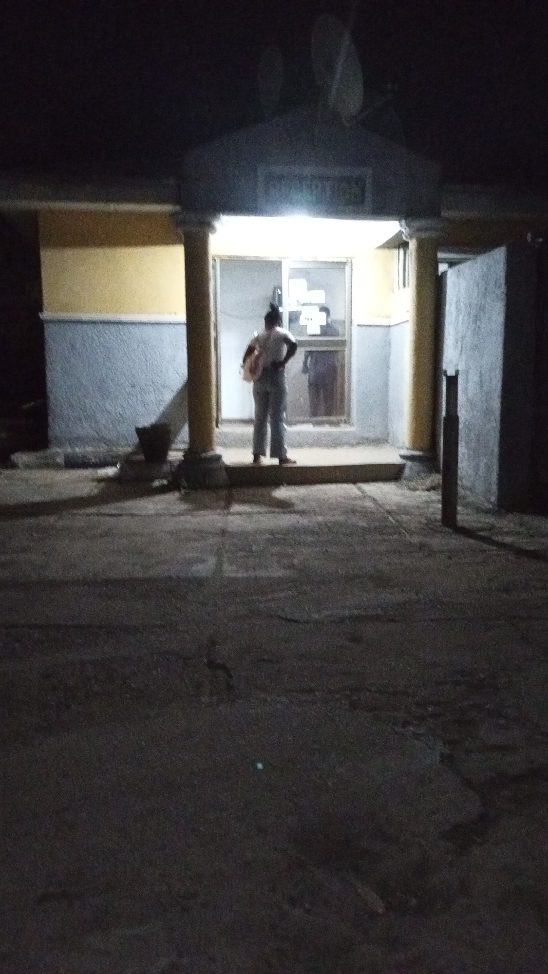 The image, captured at night in Adankolo, Nigeria, depicts an individual standing on a raised platform at the brightly lit entrance of a building. The person, viewed from behind, appears to be an adult, possibly female, with hair tied up. They are dressed in a light-colored top and light-colored trousers, with a bag or backpack slung over their left shoulder. Their hands are placed on their hips as they face the building's entrance, which features glass doors or large windows. The illuminated interior is visible, and a reflection of the person can be faintly seen in the glass. Inside, several white objects, one resembling a cross shape, are affixed to the interior wall.
Above the entrance, on the building's facade, a sign with faint, illuminated text reads "INFORMATION". Two large satellite dishes are mounted on the roof or overhang above the sign. The building itself is painted with a yellow upper section and a grey or blue lower section, supported by two prominent columns.
To the left of the person, near one of the columns, a dark, cylindrical object, possibly a planter or bin, is situated. On the right side of the frame, a dark, slender post or bollard stands vertically. The paved ground leading to the entrance is mostly shrouded in shadow, with only the immediate area in front of the doorway illuminated. The overall darkness of the scene highlights the artificial light emanating from the building. The person appears to be either waiting or observing the activity within the establishment.