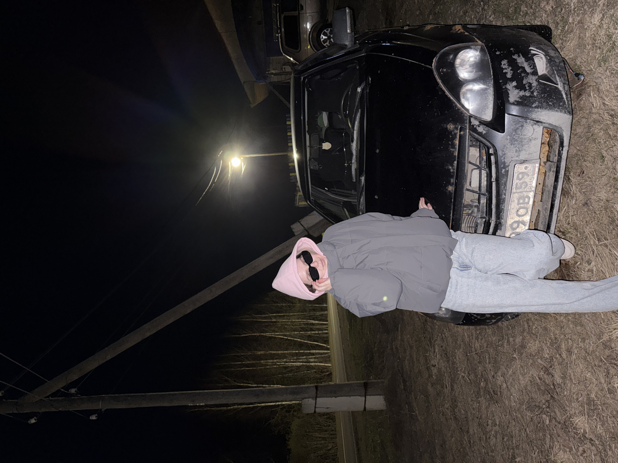 This nighttime image, likely set in Zhigalki, Russia, features a person leaning against a dark-colored car. The individual wears a gray puffer jacket over a pink hooded sweatshirt with the hood up, light blue jeans, and dark sunglasses. Their posture appears casual, with one hand resting on the car's hood.
The car, a sedan, shows significant signs of wear or damage, with extensive paint peeling or rust on its front bumper and hood. A Russian license plate, "X990OB 59 RUS," is visible on the front, with the "59" code indicating the Perm Krai region. The scene is illuminated by a bright street lamp positioned on a wooden utility pole, with other poles and wires stretching across the dark sky. In the background, beyond the main car, another vehicle, possibly a van or truck, is partially visible. Bare trees suggest an autumn or winter season. The ground is unpaved, consisting of dry grass or dirt, indicative of a rural or semi-rural roadside setting. The low light conditions and artificial illumination create a stark contrast.