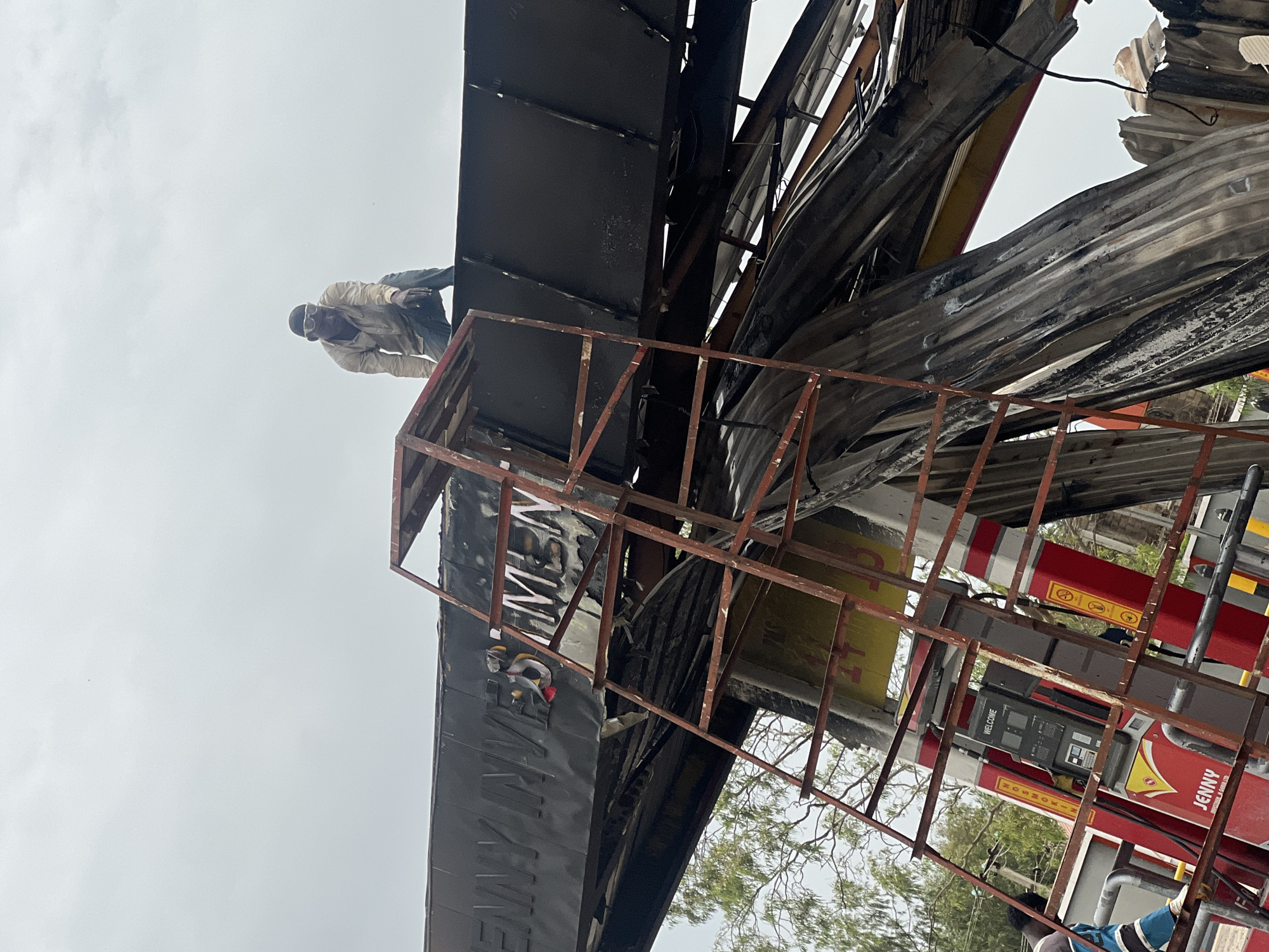 A man wearing light-colored clothing and eye protection stands on the edge of a damaged structure. The structure appears to be the burnt remains of a canopy or sign, possibly from a Jenny fuel station, based on the red and yellow colors and visible word "JENNY" on the fuel pumps. The location is Gura Top, Nigeria. The canopy is severely damaged, with twisted and burnt metal. A red metal frame is in the foreground. The sky is overcast.