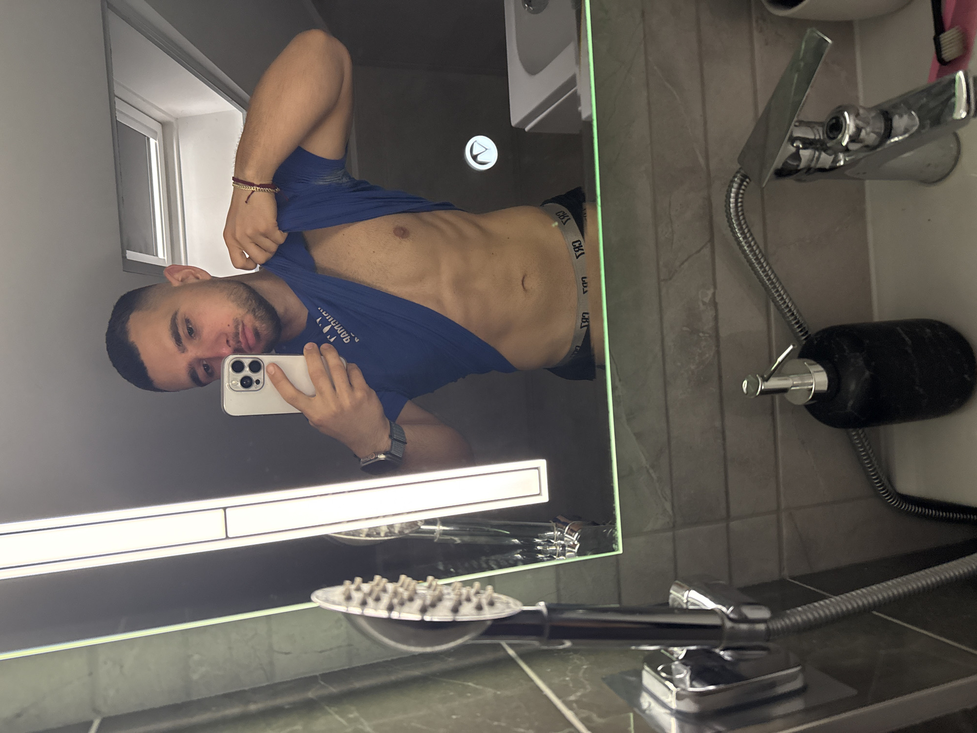 A man is taking a selfie in a bathroom mirror. He is shirtless, wearing underwear and a blue t-shirt, which he is partially lifting. The bathroom contains a sink, a shower head, and gray tiling. The location is not possible to determine from the image, beyond it being a bathroom.