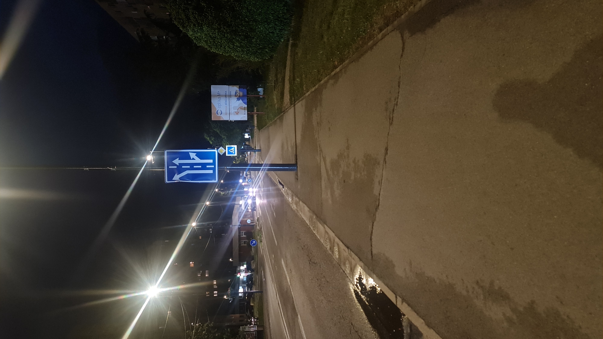 The image depicts a street at night in an urban environment. A blue traffic sign indicating lane directions is prominently visible. Streetlights and car headlights provide illumination. A sidewalk runs along the right side of the street, showing signs of recent rain or dampness. A billboard and some greenery are seen in the background. The location is unknown.