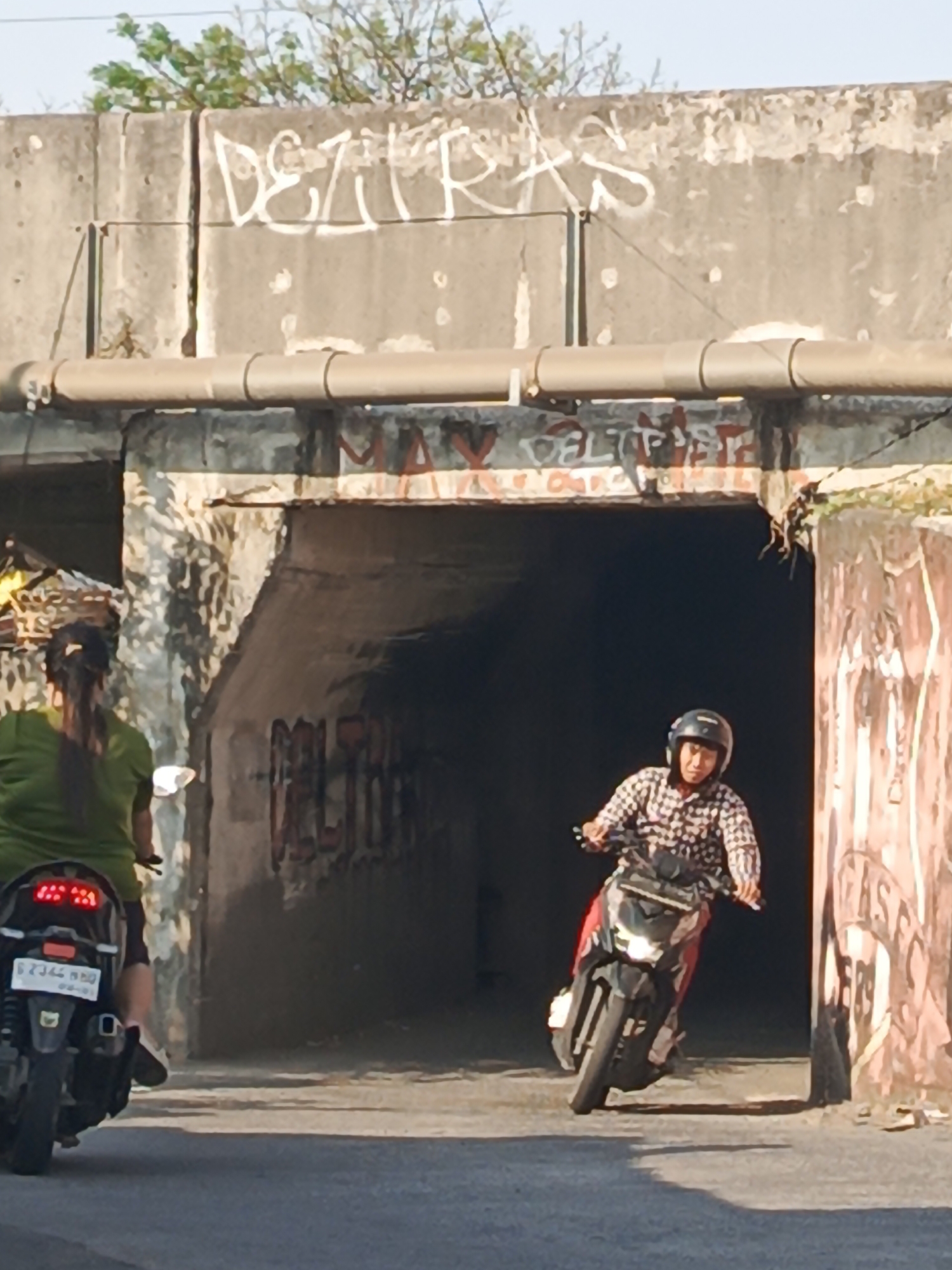 In Kludan, Indonesia, two individuals on motorcycles are seen near a concrete underpass. The underpass is adorned with graffiti, including the word "DEZIRAS" in white on the upper concrete structure, and red graffiti on the walls within the underpass. A large pipe runs horizontally along the top of the underpass.
In the foreground and to the left, a person wearing a green shirt rides a black motorcycle. The motorcycle's rear features a license plate with the characters "B 2346 BBO".
In the mid-ground and center, a person wearing a checkered shirt and a black helmet rides a black and red motorcycle. This individual is partially obscured by the underpass opening and appears to be moving towards the viewer. The overall scene depicts an urban environment with signs of graffiti art and everyday transportation.