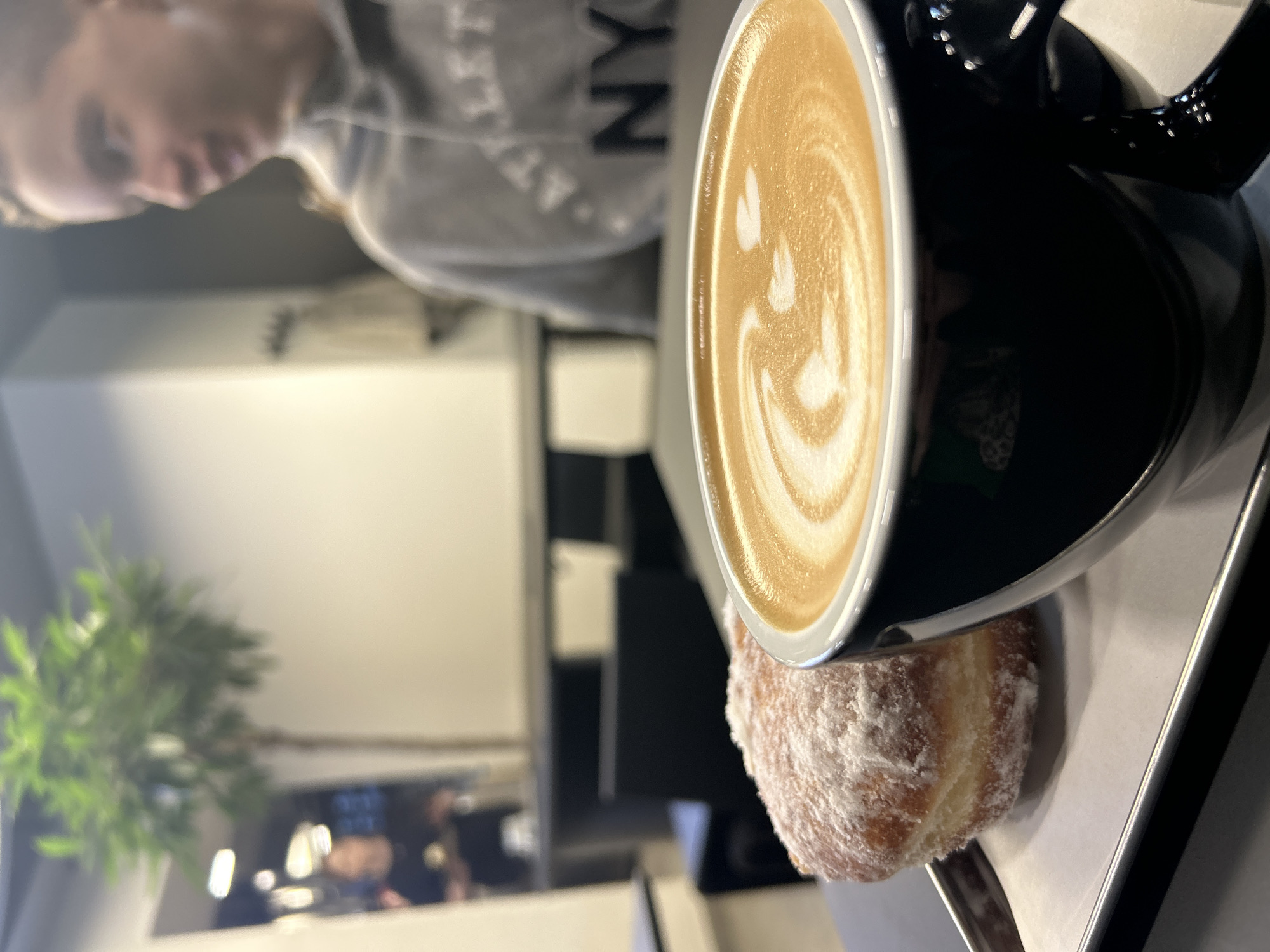 A black coffee mug filled with latte art rests on a silver tray next to a powdered sugar-covered donut. A person wearing a gray hoodie with "NYC" printed on it sits in the background, slightly out of focus. In the deeper background, there is a green potted plant and indistinct figures near a window. The scene appears to be indoors, potentially in a cafe setting. No specific details indicate the city of Izhevsk, Russia.