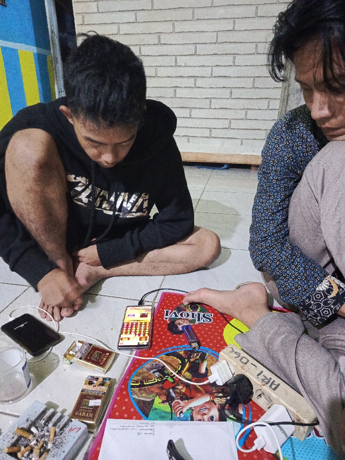 Two young men are seated on a tiled floor in what appears to be an indoor setting in Salatiga, Indonesia. The person on the left wears a black hoodie with "ZELDA" written on the front and is looking downwards. The other person, on the right, wears a blue patterned long-sleeved shirt and light-colored pants, also looking down, with only their upper body and legs visible.
In front of them, scattered on the floor, is an array of personal items. Two smartphones are visible, both connected to charging cables. One phone, centrally placed, displays what appears to be a mobile game interface with multiple colored blocks. Next to it are two packs of "GARAM" brand cigarettes, one open and one closed, along with an ashtray filled with cigarette butts. The charging cables lead to a multi-socket power strip, which is a piece of wood with several power outlets, labeled with the text "ARI-060". This charging setup rests on a red mat or book that features various cartoon-like images and the text "Shovi".
The activity centers around the use of mobile phones, possibly for gaming, while smoking. The scene depicts a casual gathering, likely in the evening or at night, suggested by the indoor artificial lighting and the light-colored brick wall in the background. The men appear focused on their respective activities. In the far left background, a blue and yellow striped object is partially visible.