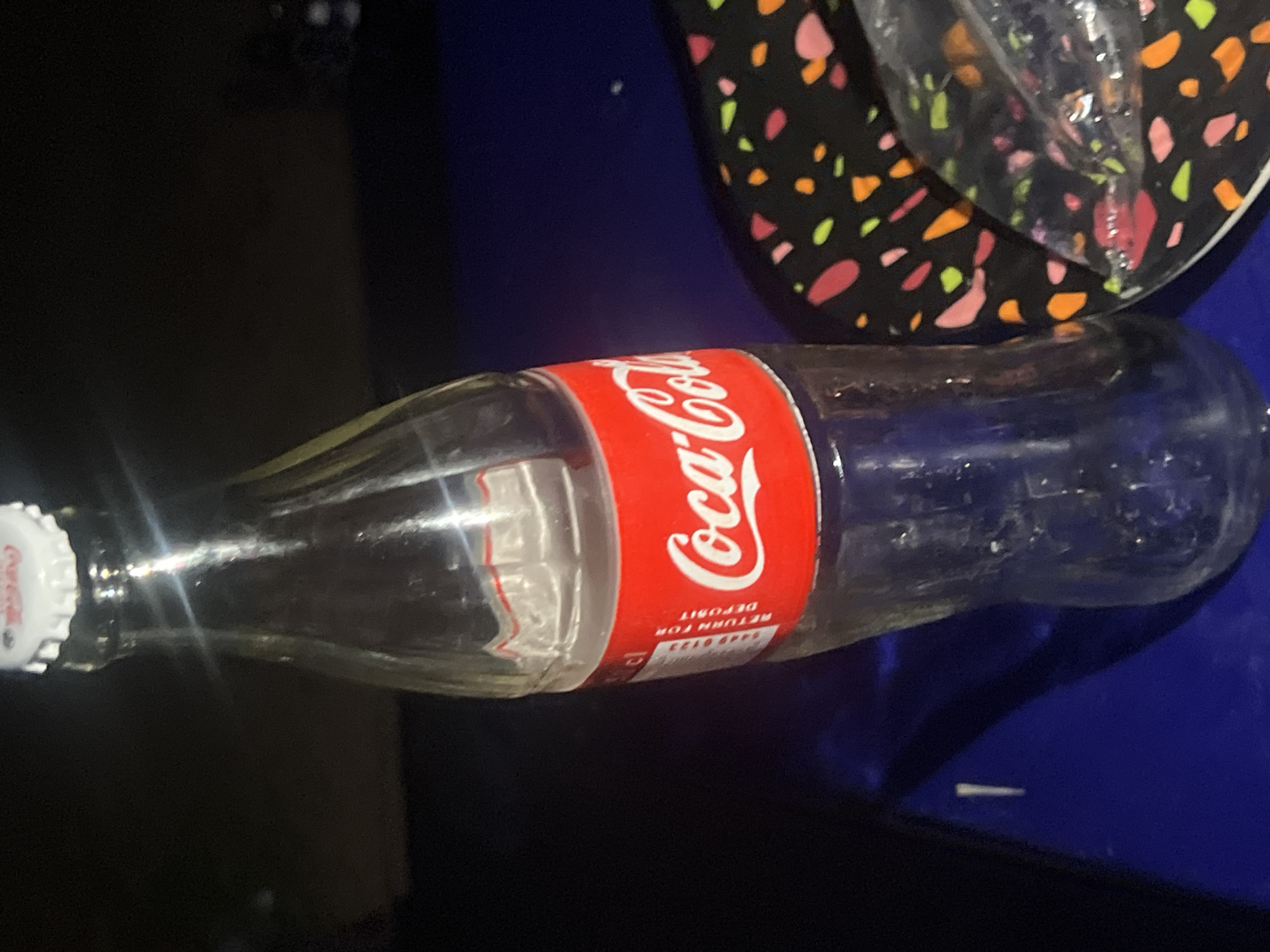 An empty Coca-Cola bottle lies horizontally on a dark surface. A colorful hat rests next to the bottle. The image provides no information about Jalingo, Nigeria.
