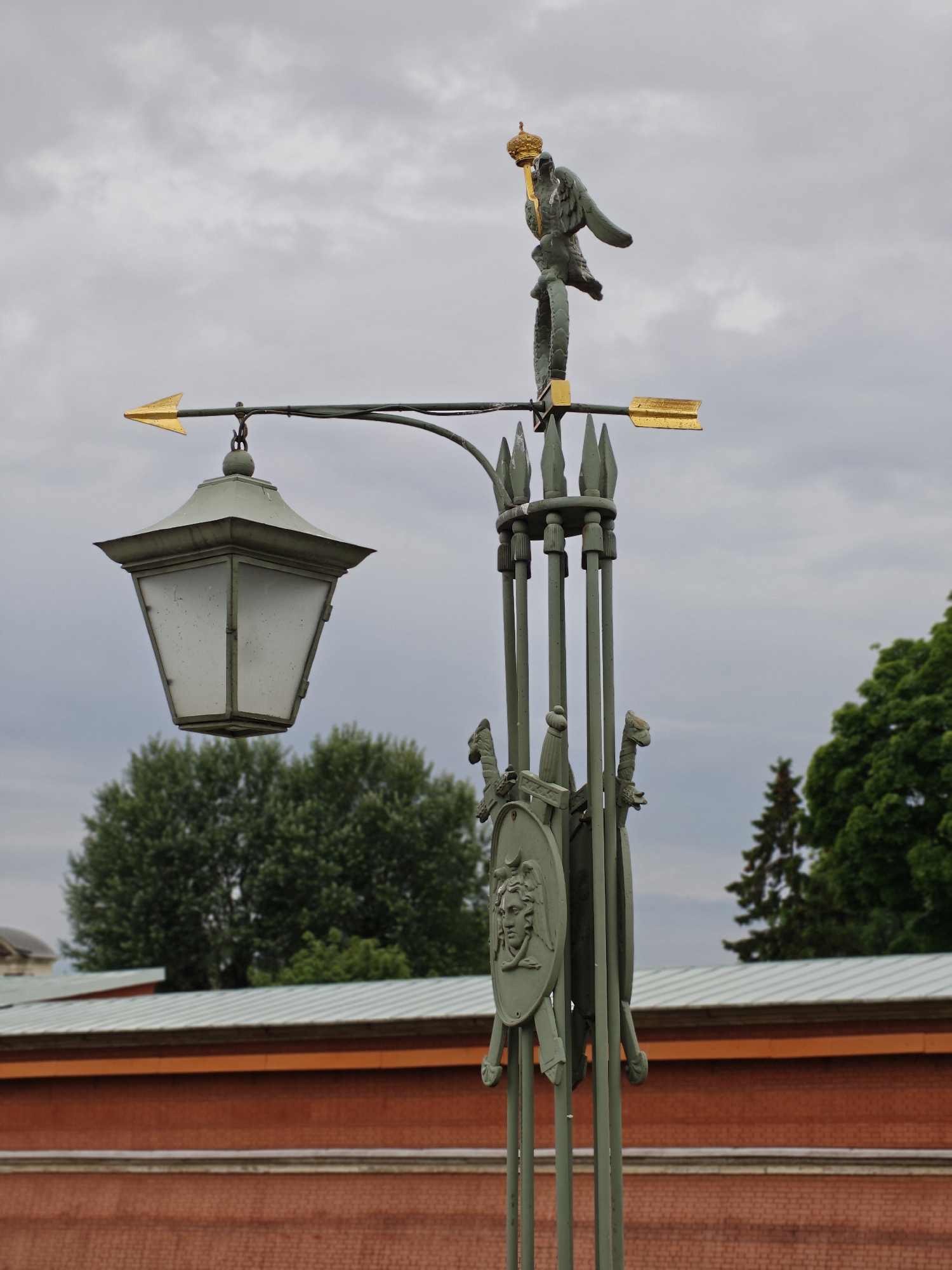 This is a medium-shot, eye-level photograph of an ornate lamppost topped with a metal eagle statue. The lamppost is made of dark green metal and is elaborately decorated with elements such as swords, shields, and what appears to be a crown on the eagle's head. A gold arrow wind vane is attached to the lamppost. Below the lamppost, a large, pale green lantern hangs from an arched arm. The background features a cloudy sky, lush green trees, and a red brick building with a light-colored roof. The building appears to be part of a historical site, possibly a palace or museum, given the architectural style and decorative elements on the lamppost. This lamppost is located in Saint Petersburg, Russia, specifically on the Peter and Paul Fortress.
